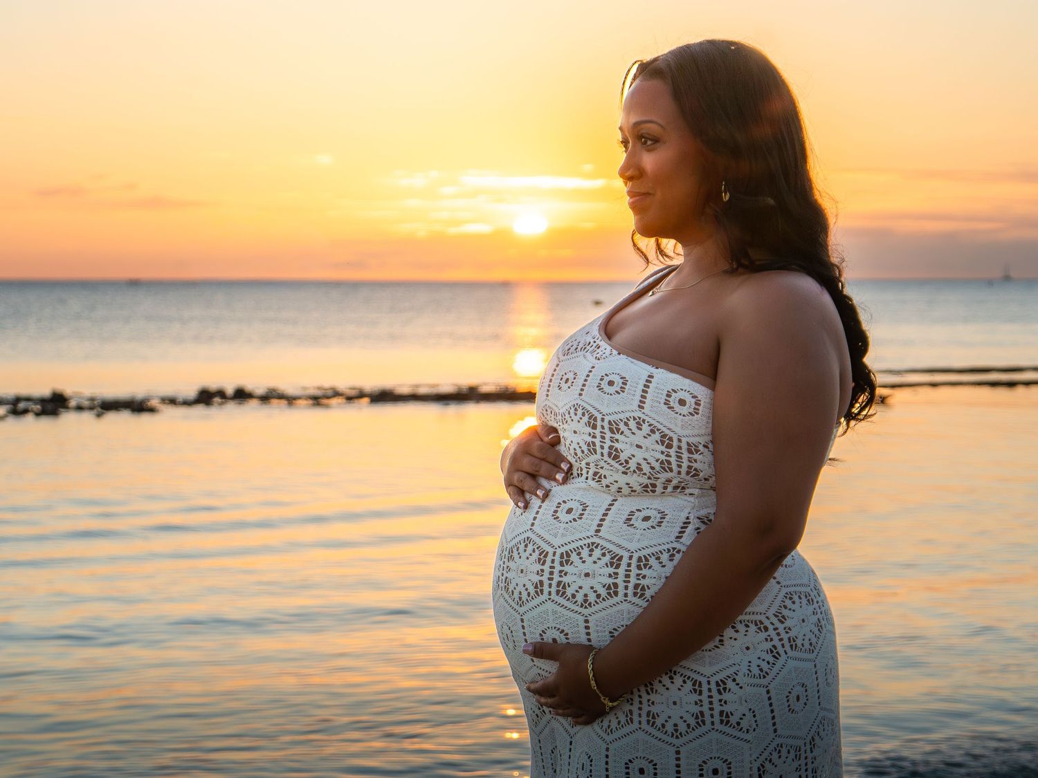 Pregnant woman in white lace dress holding her belly at sunset by the ocean.