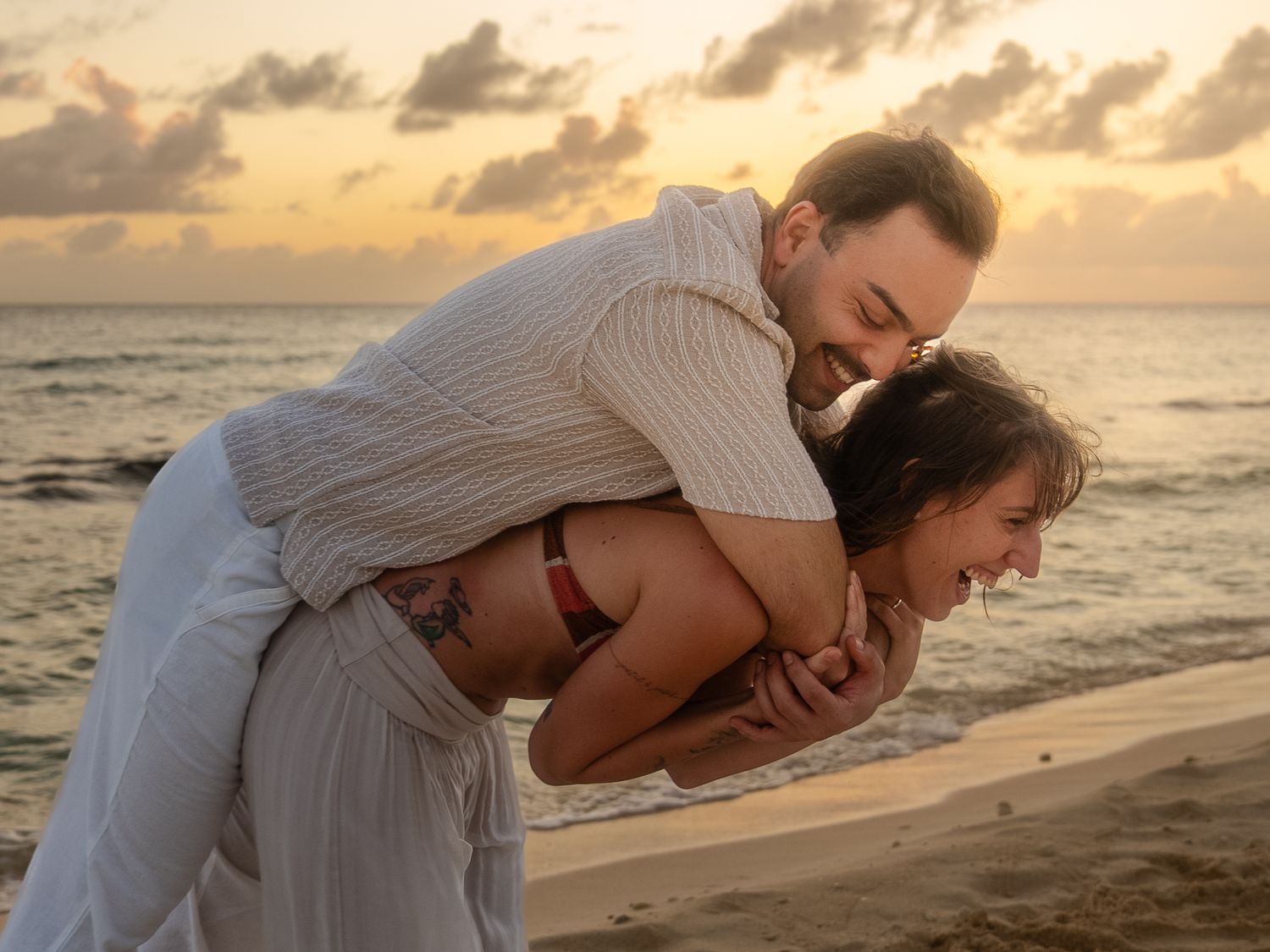 Romantic couple laughing on sandy beach at sunset, man lifting woman piggyback.