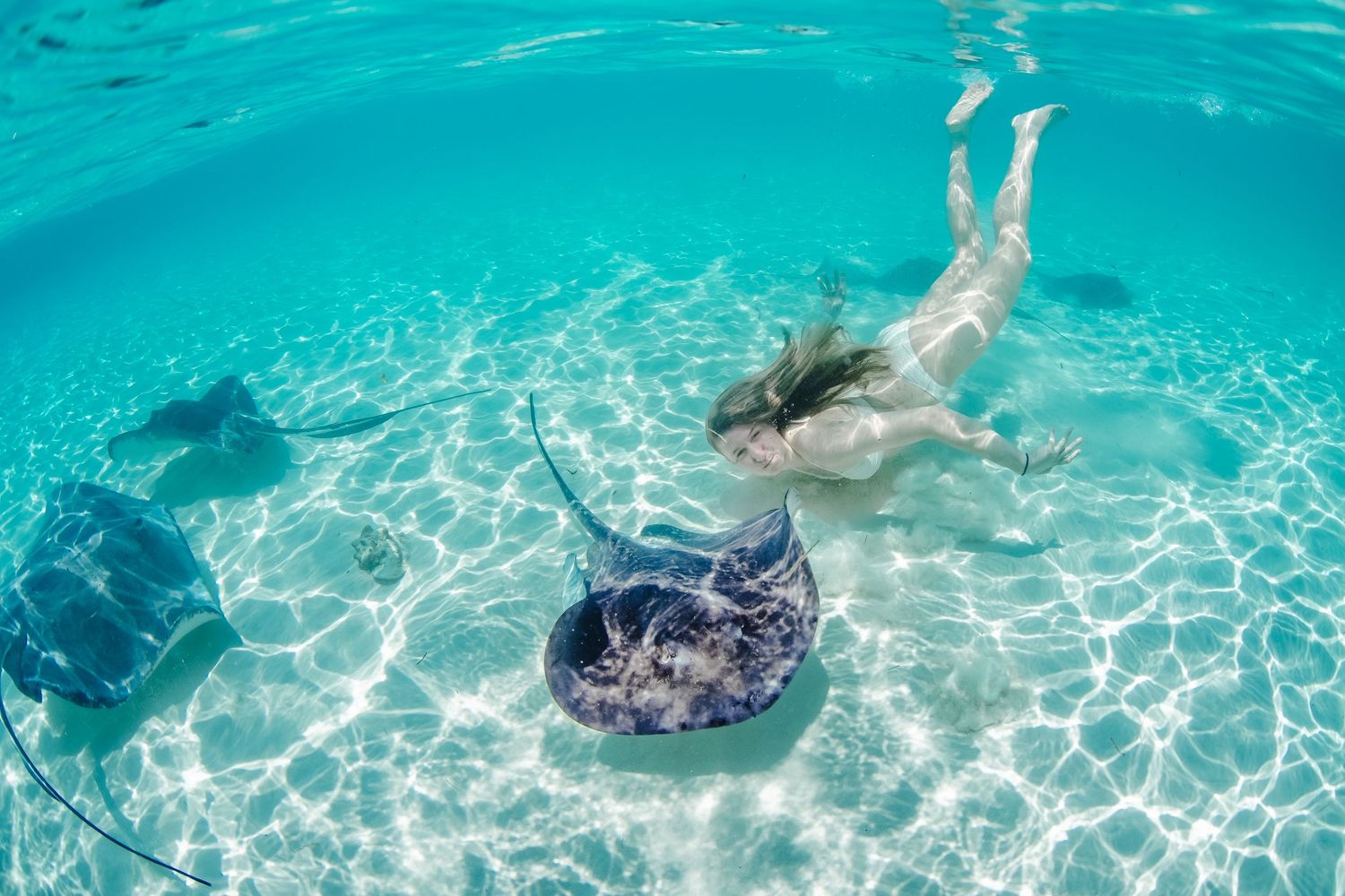 Underwater swimmer gliding over white sandy ocean floor with rippling sunlight patterns.