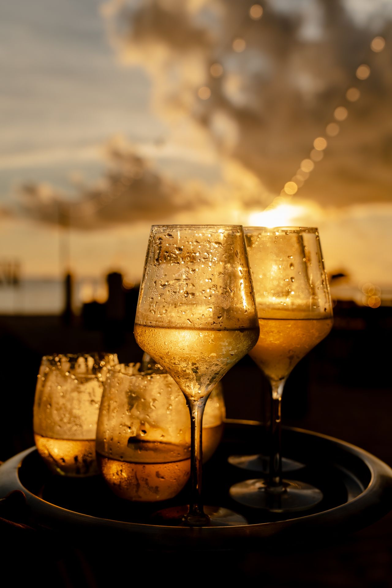 Wine glasses filled with golden liquid catch the light of a dramatic sunset.