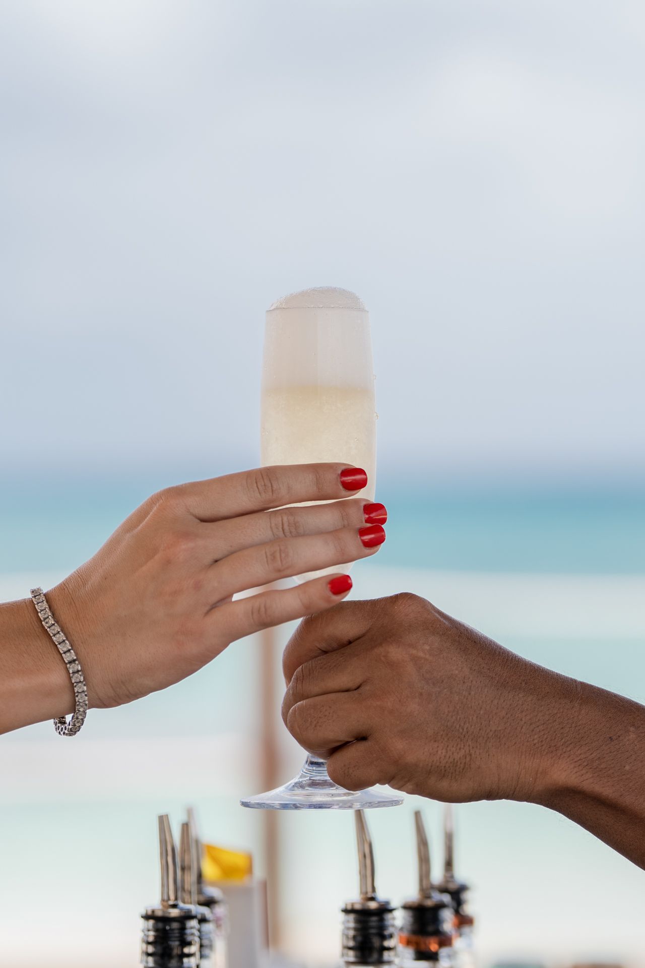 Hands holding frosted cocktail glasses against a tropical ocean backdrop.
