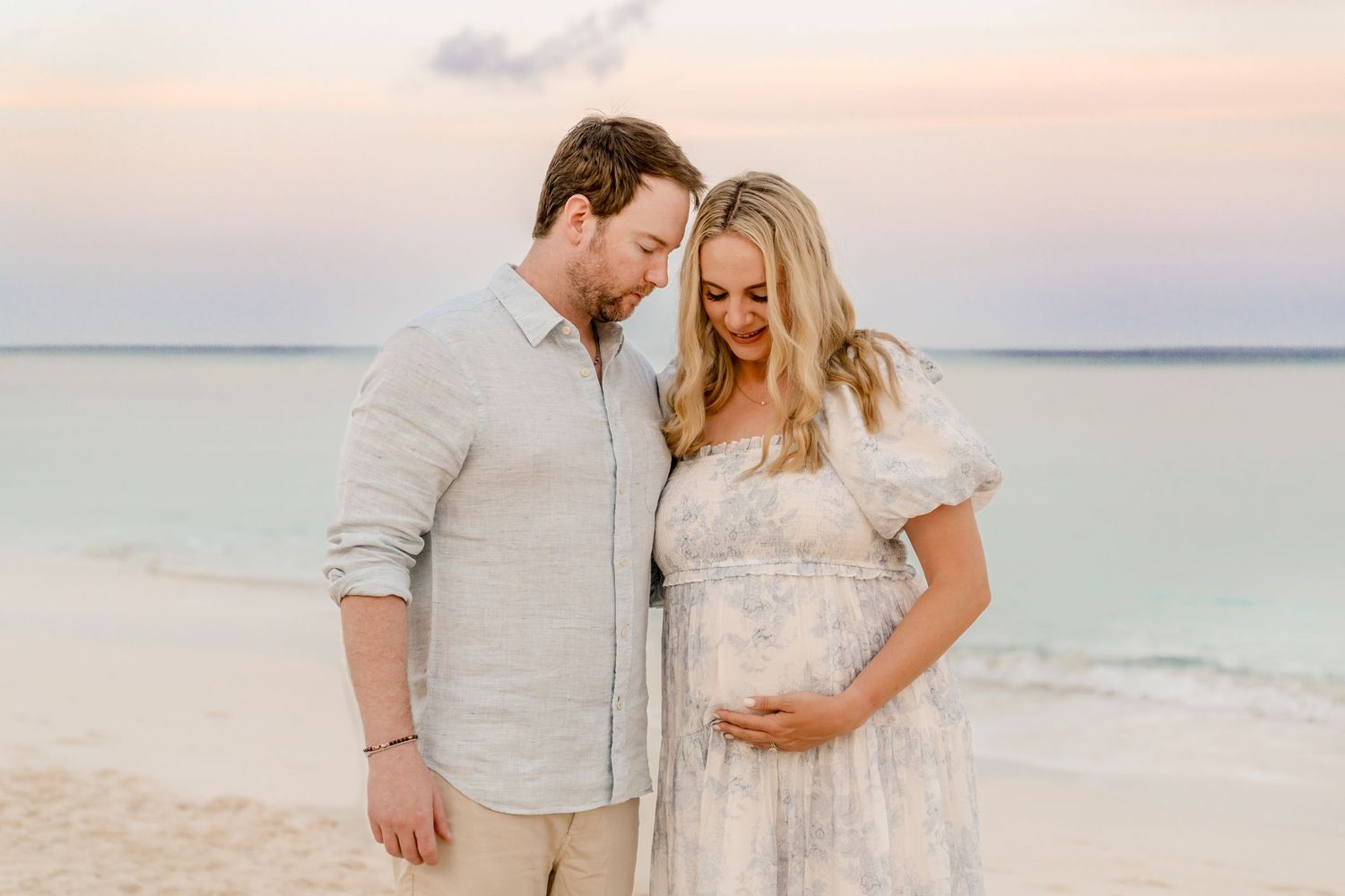 Intimate maternity beach photo of couple in light colored clothing at dusk.