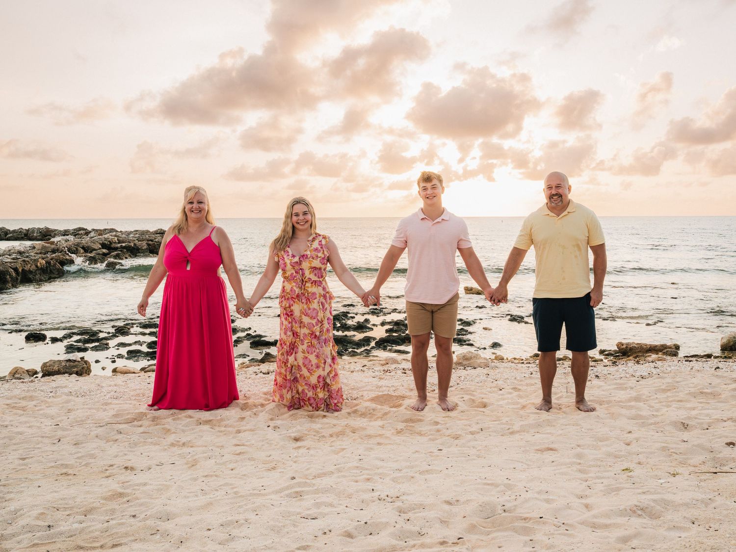 Family of four holding hands on a sandy beach at sunset with rocky shoreline and pastel sky.