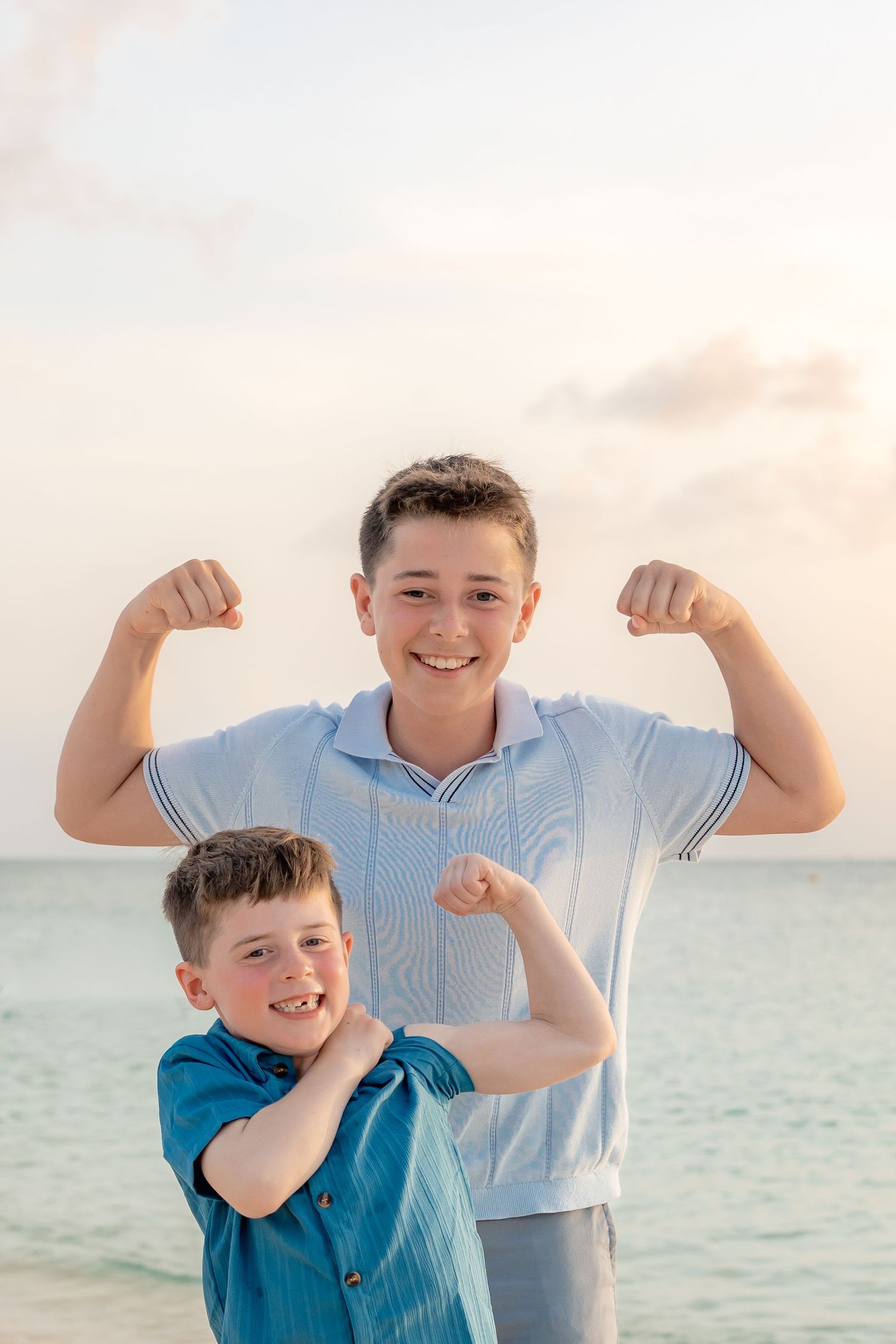 Two boys flexing muscles on a beach at sunset, smiling confidently in casual shirts with ocean backdrop.