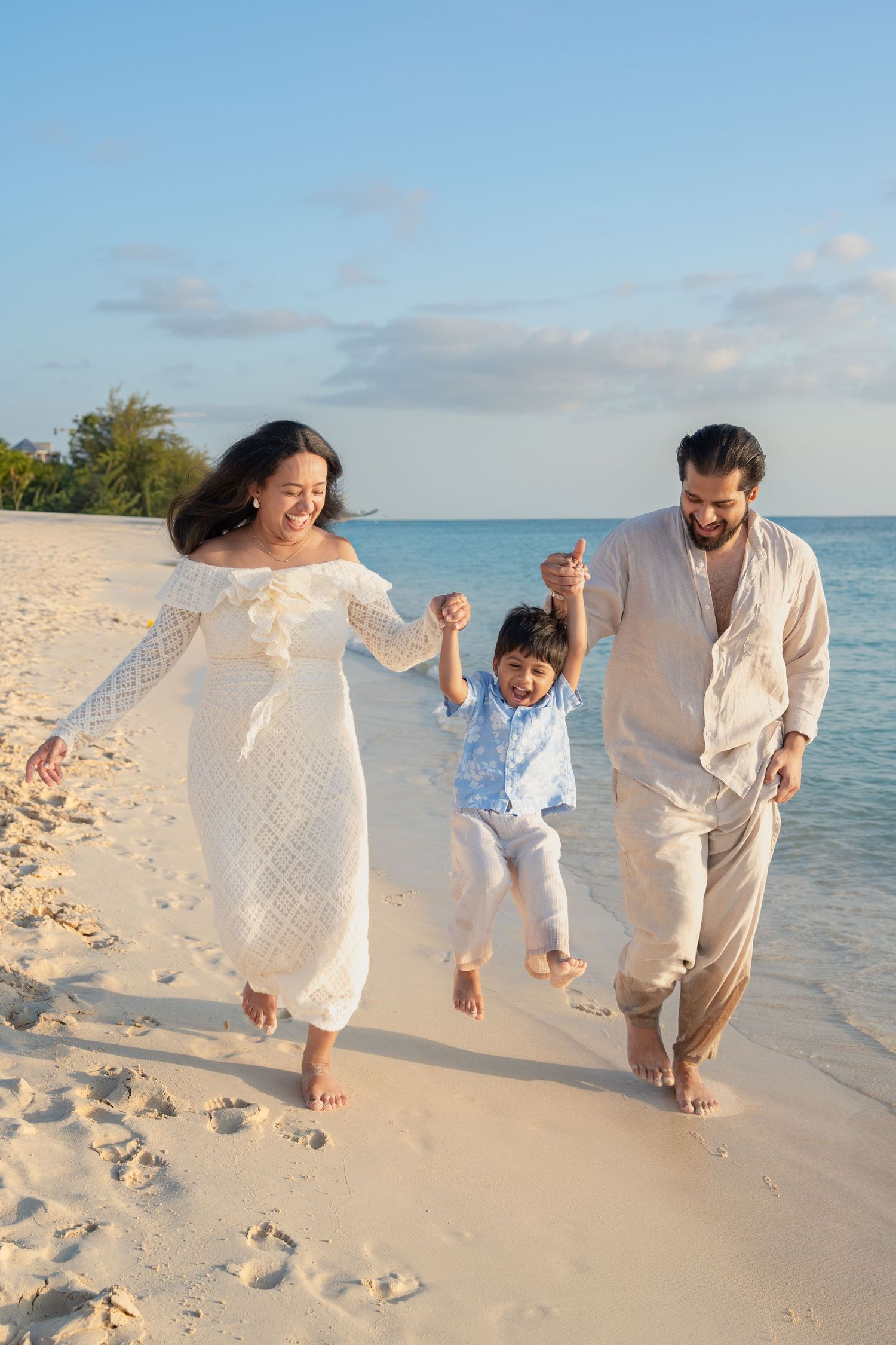Happy family with young child swinging between parents on a tropical white sand beach at sunset.