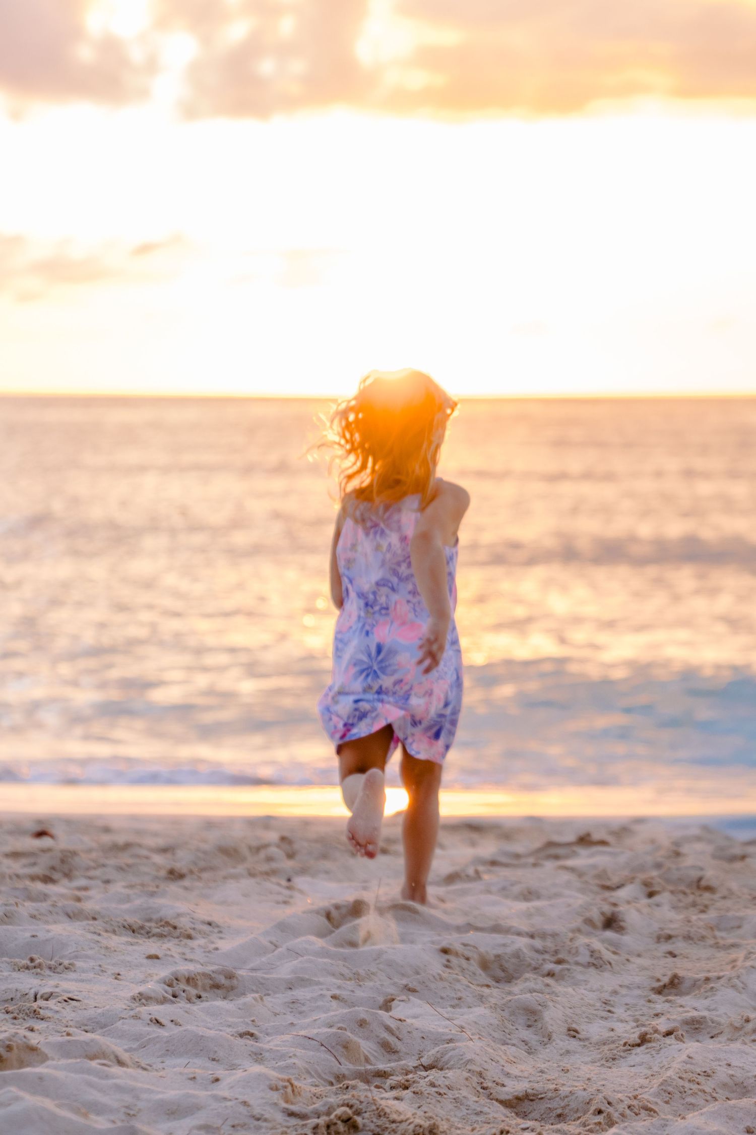 A person in a floral dress walks along a sandy beach at sunset with warm golden light reflecting off the ocean.