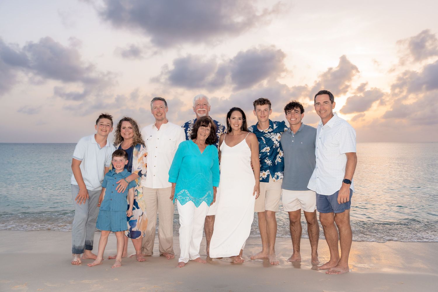 Family group portrait on a tropical beach at sunset, wearing coordinated outfits in white and blue tones.
