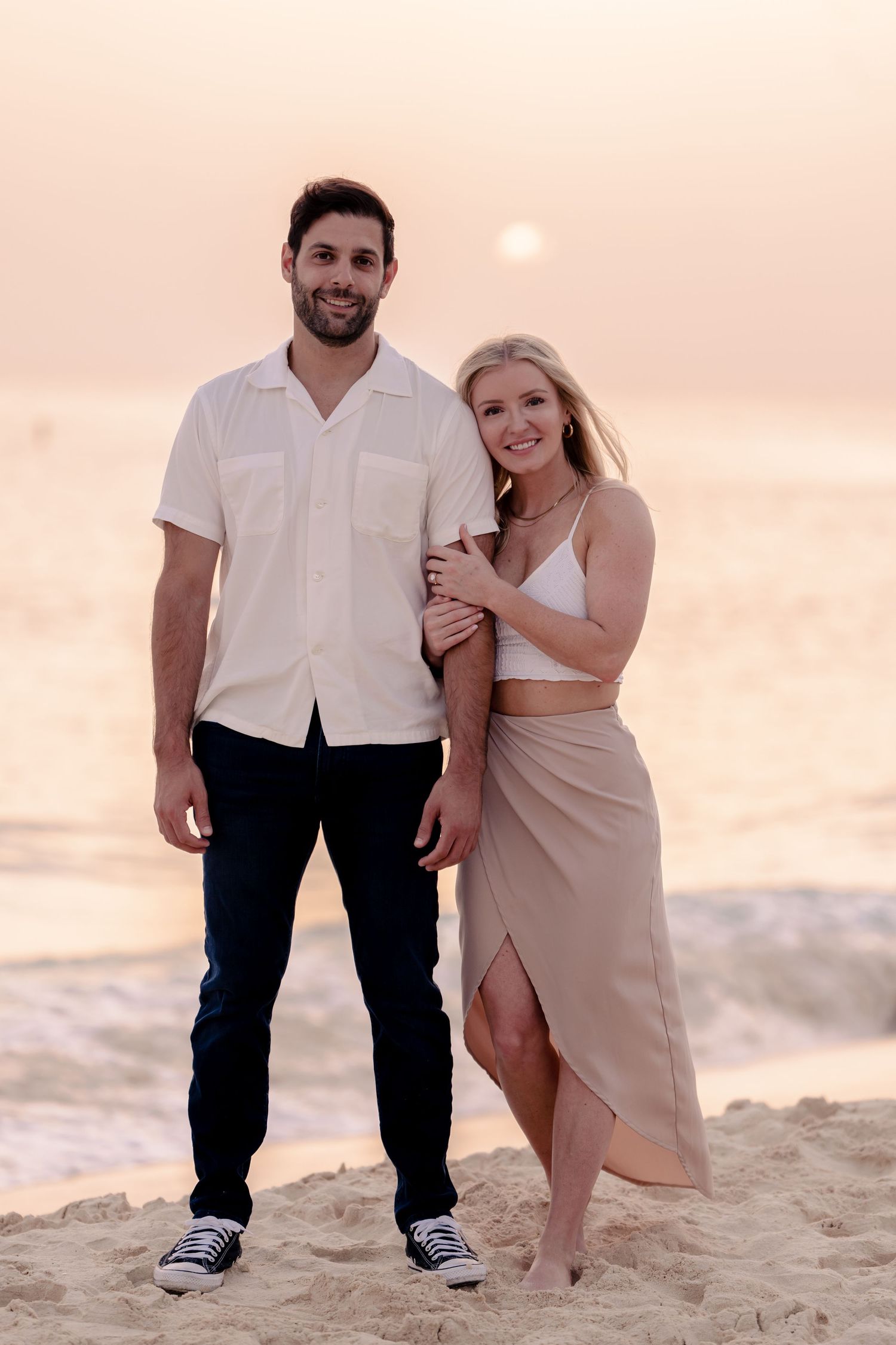 Couple poses intimately on sandy beach during sunset wearing casual white and beige outfits.