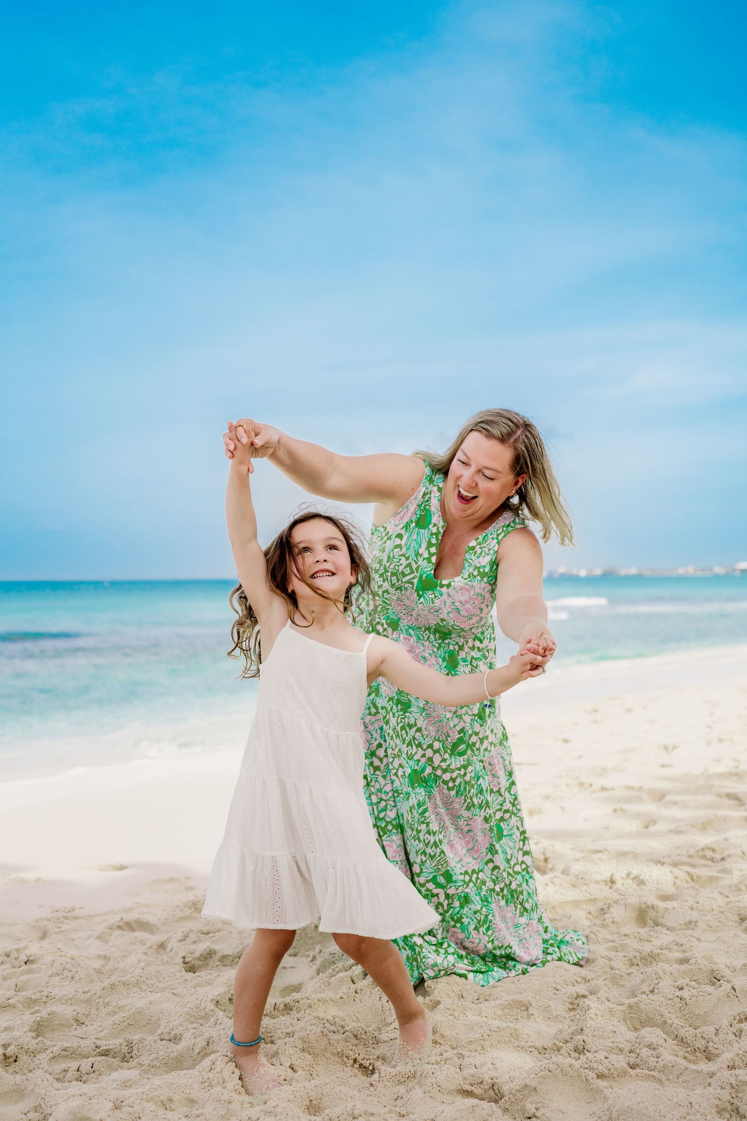 Two people in flowy dresses dancing joyfully on a tropical beach with bright blue waters.