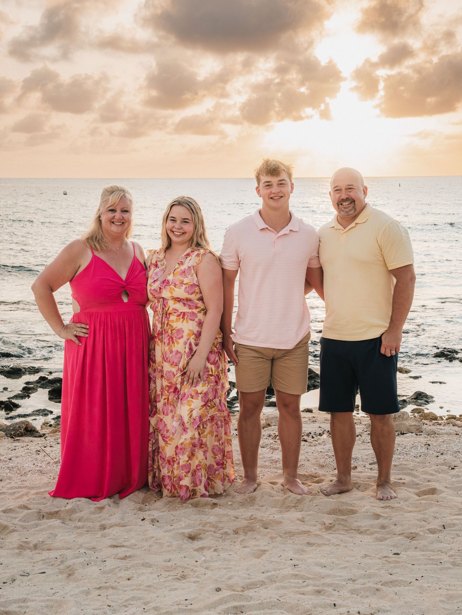 Family of four poses on sandy beach at sunset, wearing pink and yellow outfits with ocean waves behind them.
