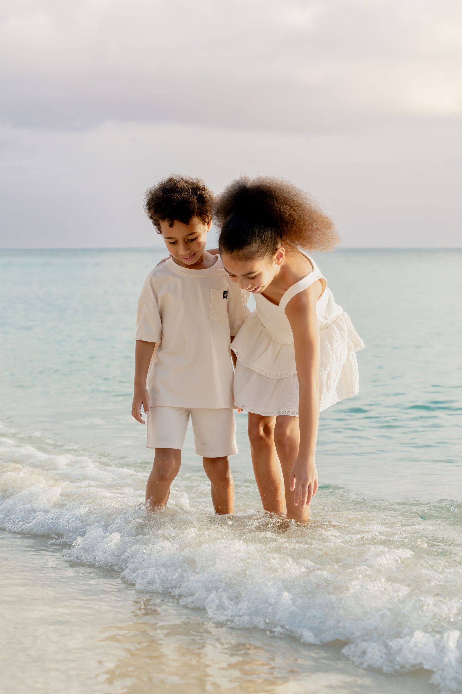 Two people in white outfits stand in gentle ocean waves on a tropical beach.