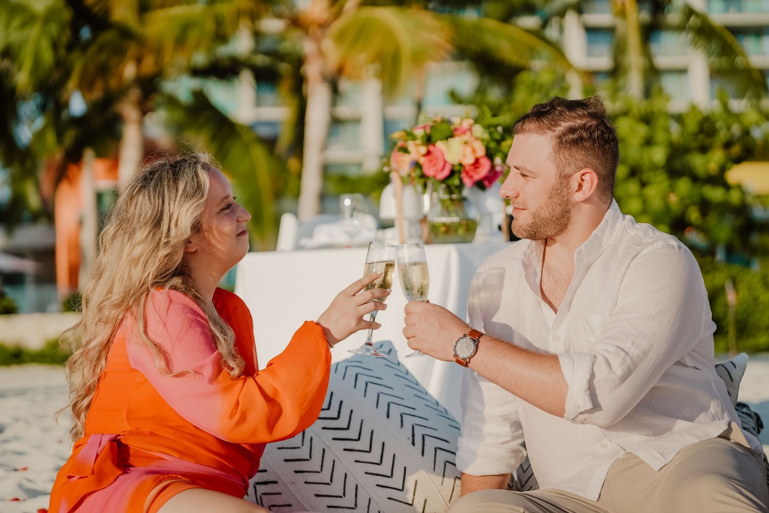 A couple sits together on a beach during sunset sharing an intimate moment in an orange and white color palette.