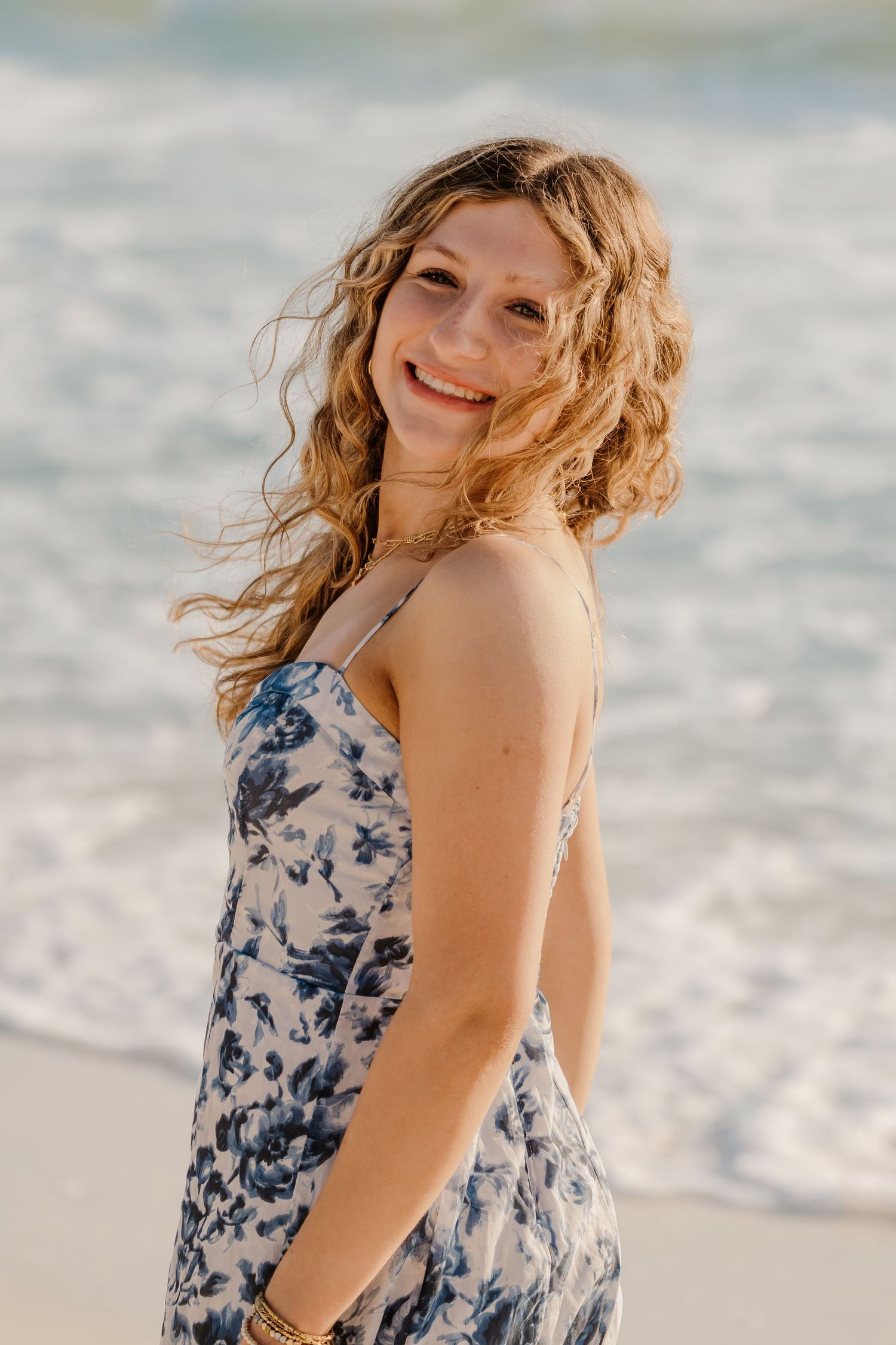 Person in blue floral dress with windswept hair poses on beach at sunset.