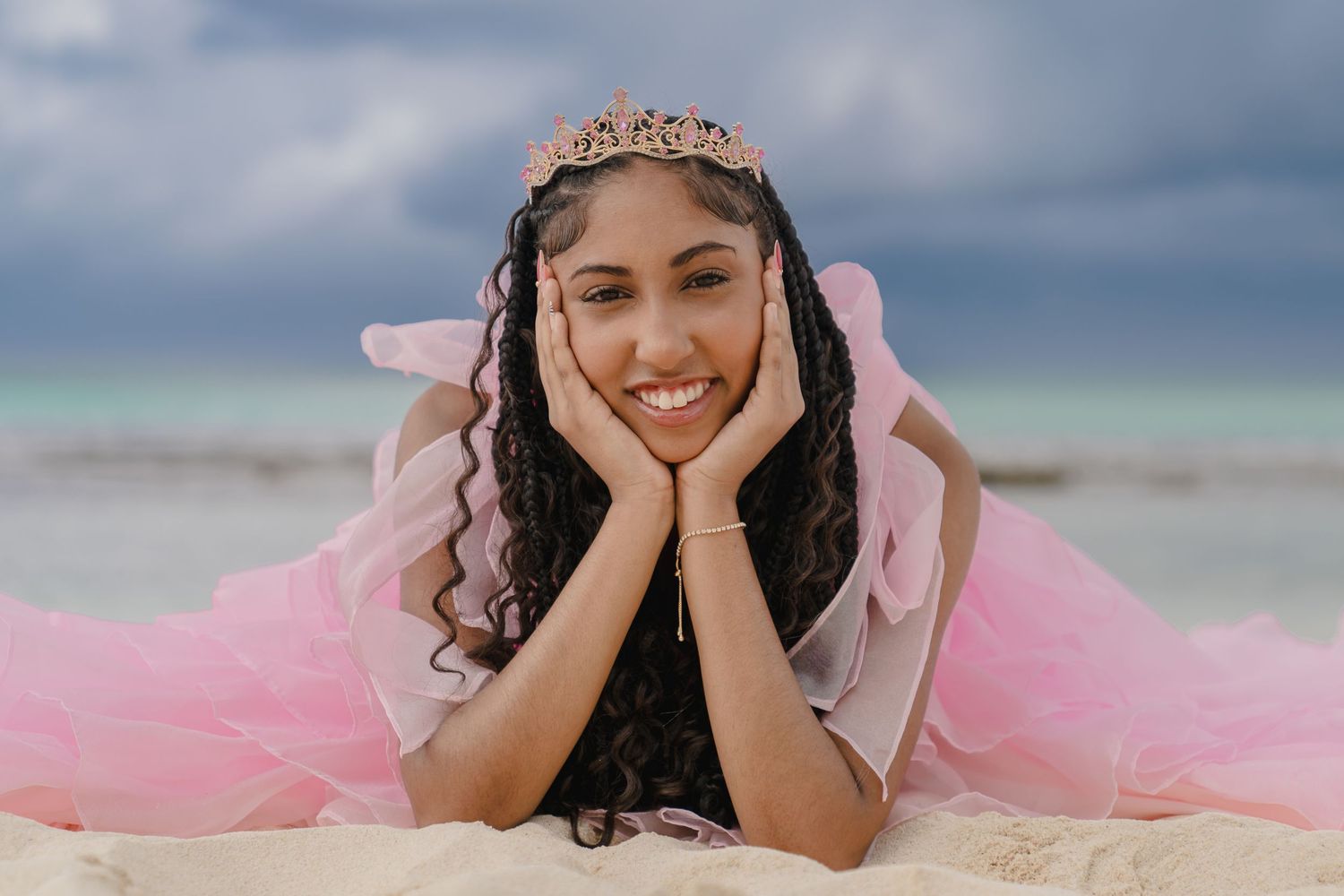 Person wearing tiara poses on sandy beach with hands framing face.
