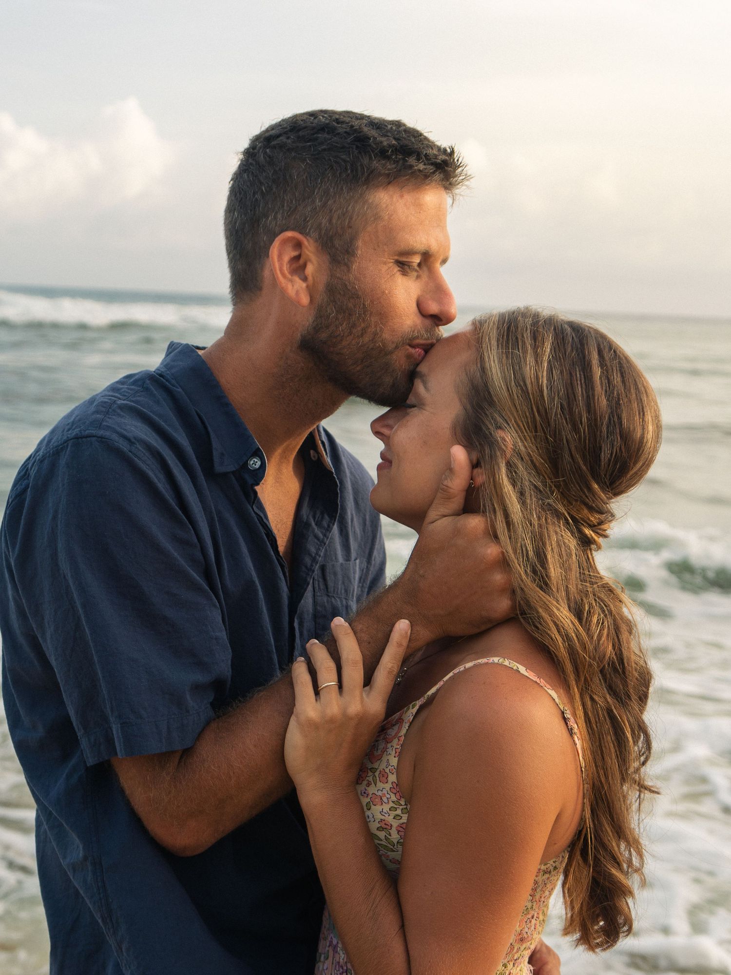 Couple sharing a romantic kiss on the beach with ocean waves in the background at sunset.