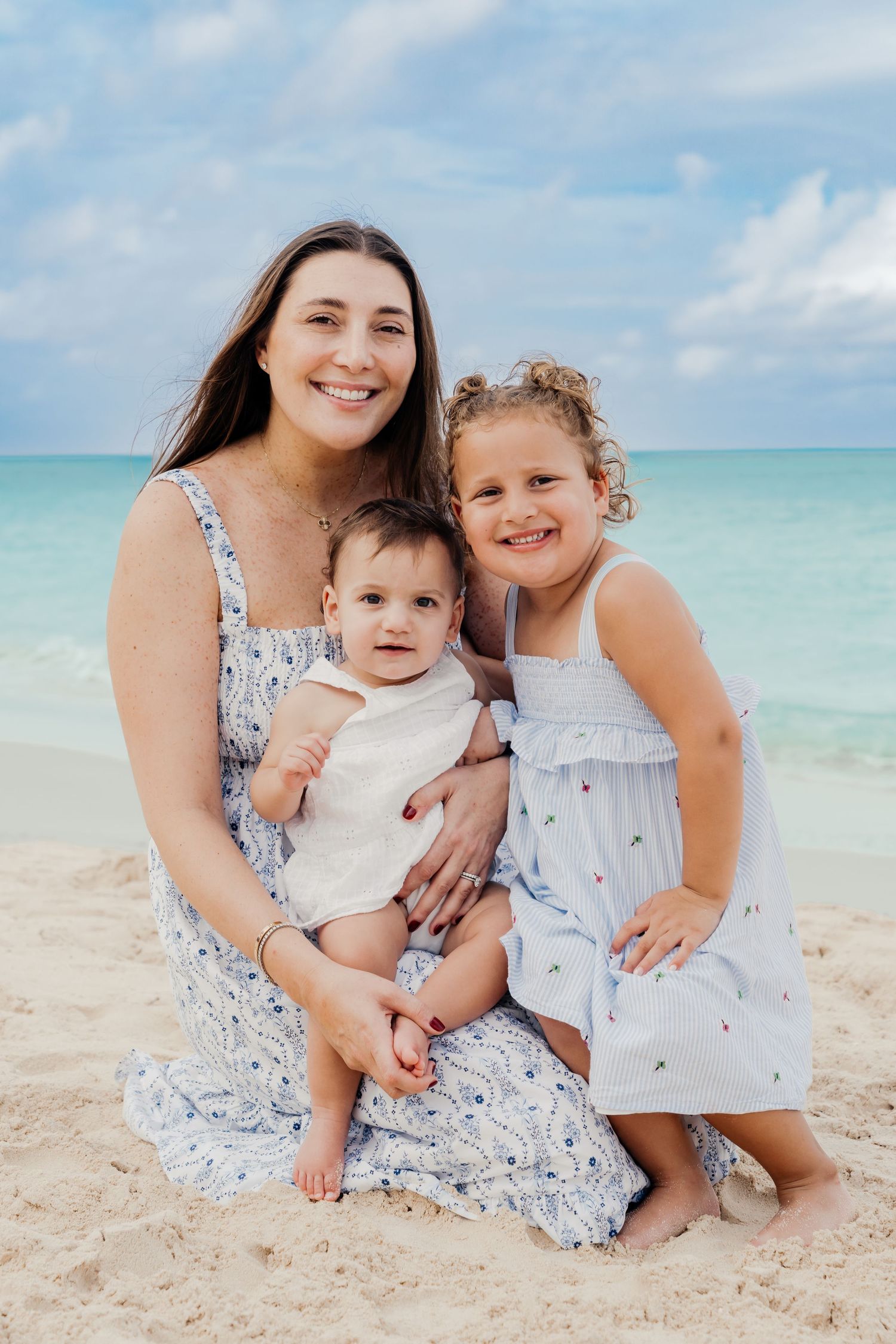 A family in matching floral dresses sits together on a tropical beach with turquoise waters and white sand in the background.