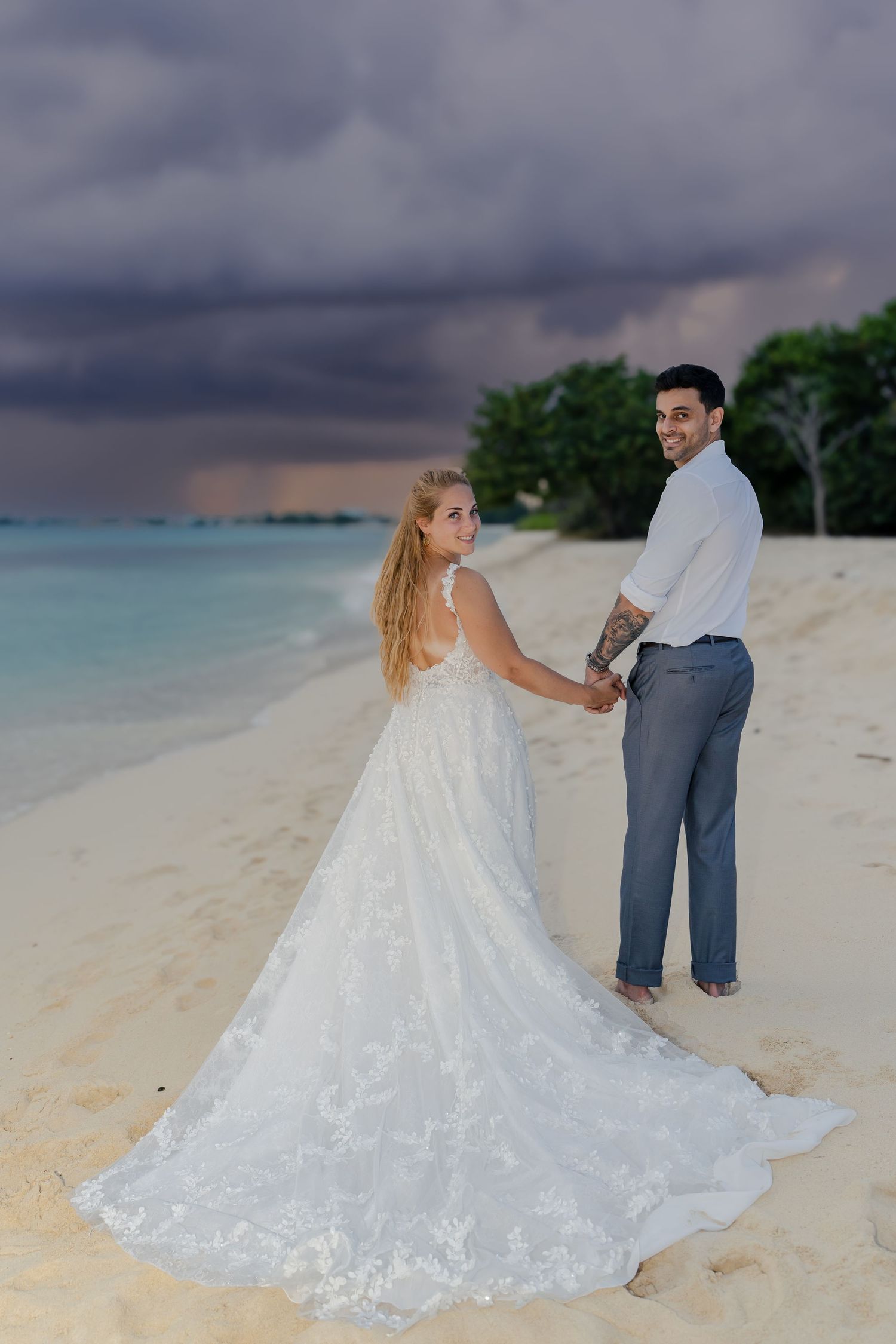 A bride's elegant lace wedding dress trails in the sand during a beach photoshoot at dusk.
