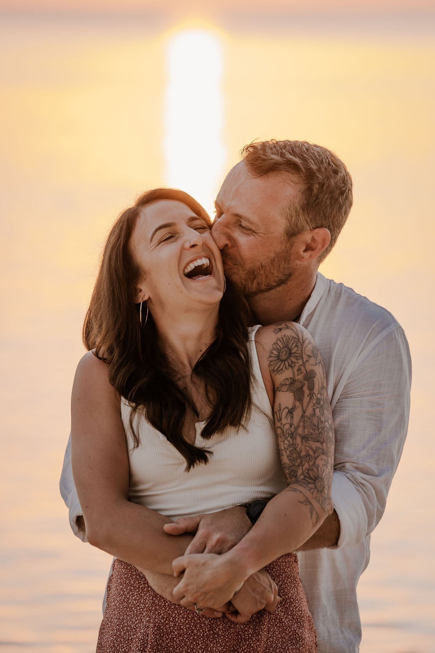 A joyful couple embracing and laughing during a sunset photoshoot by the water.