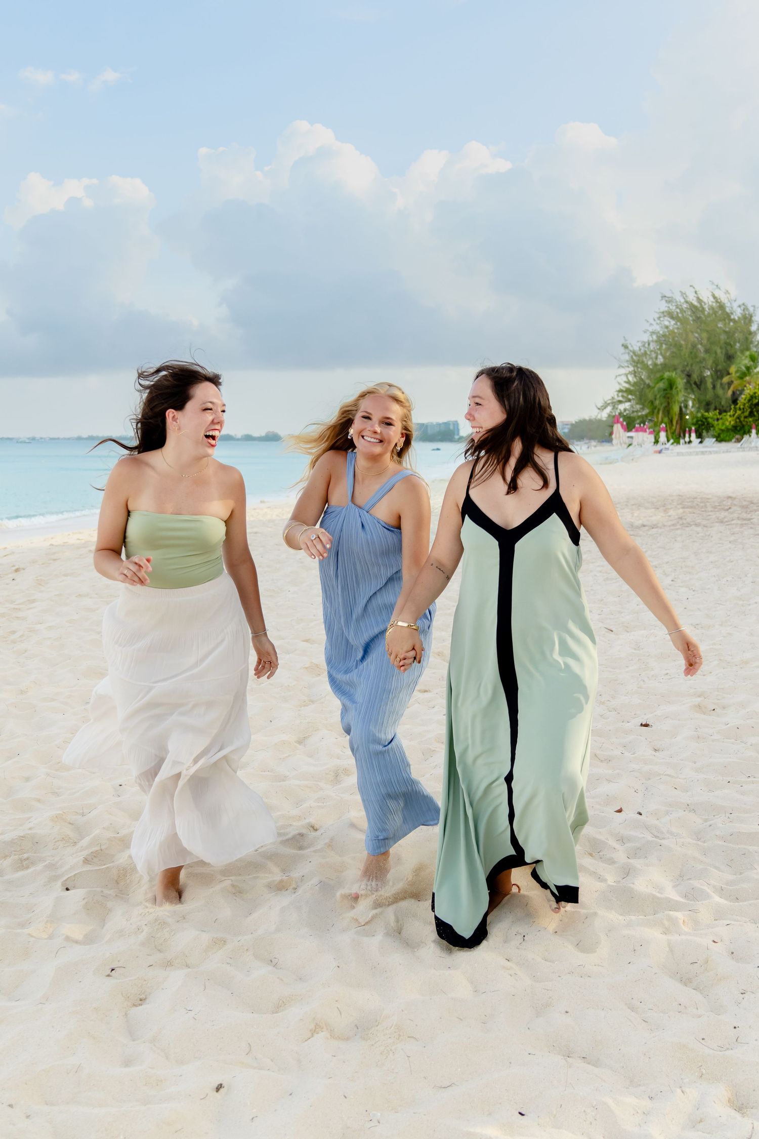 Friends in flowing dresses run and laugh together on a white sand beach.