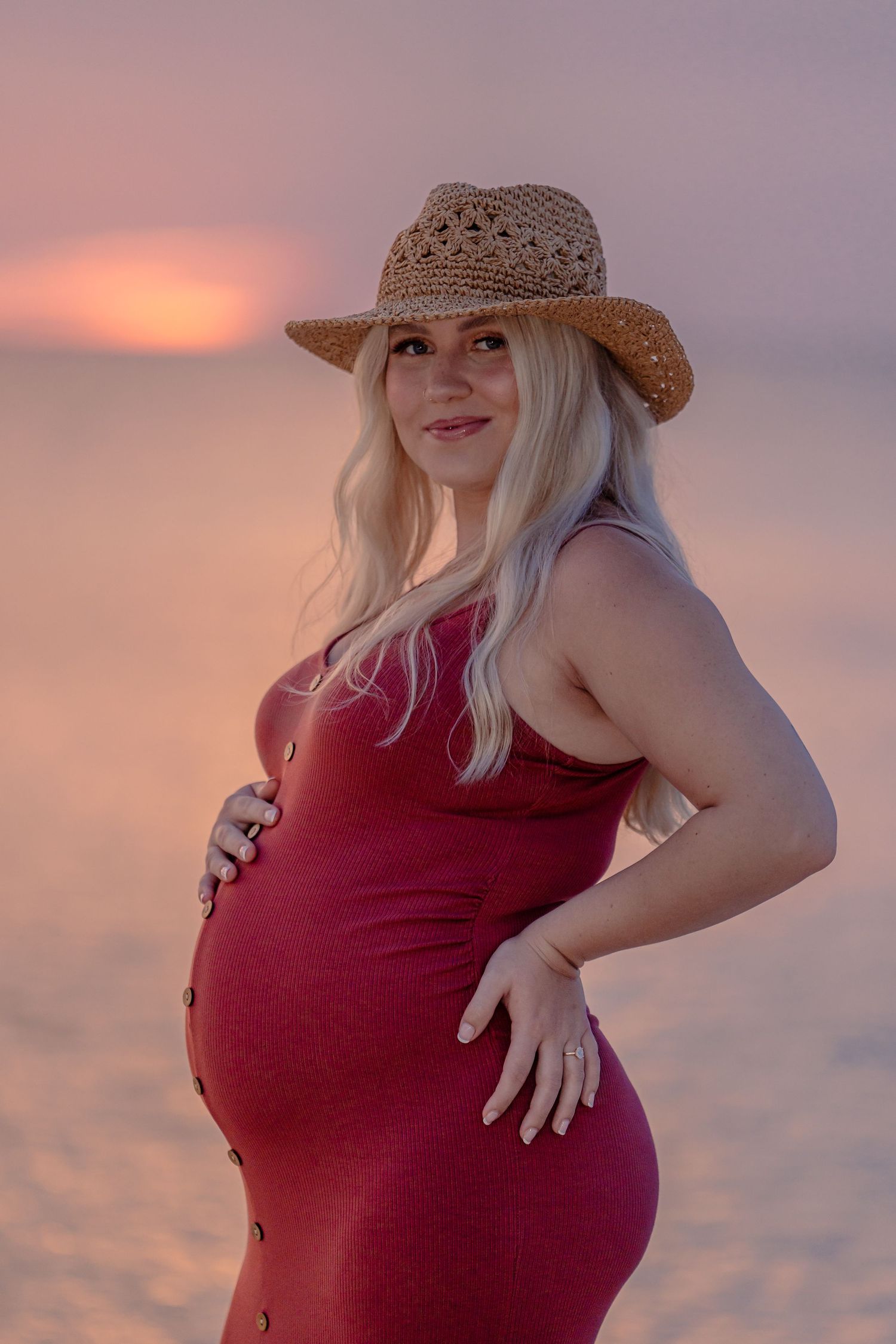 Sunset maternity portrait with straw hat and burgundy dress against pink sky.