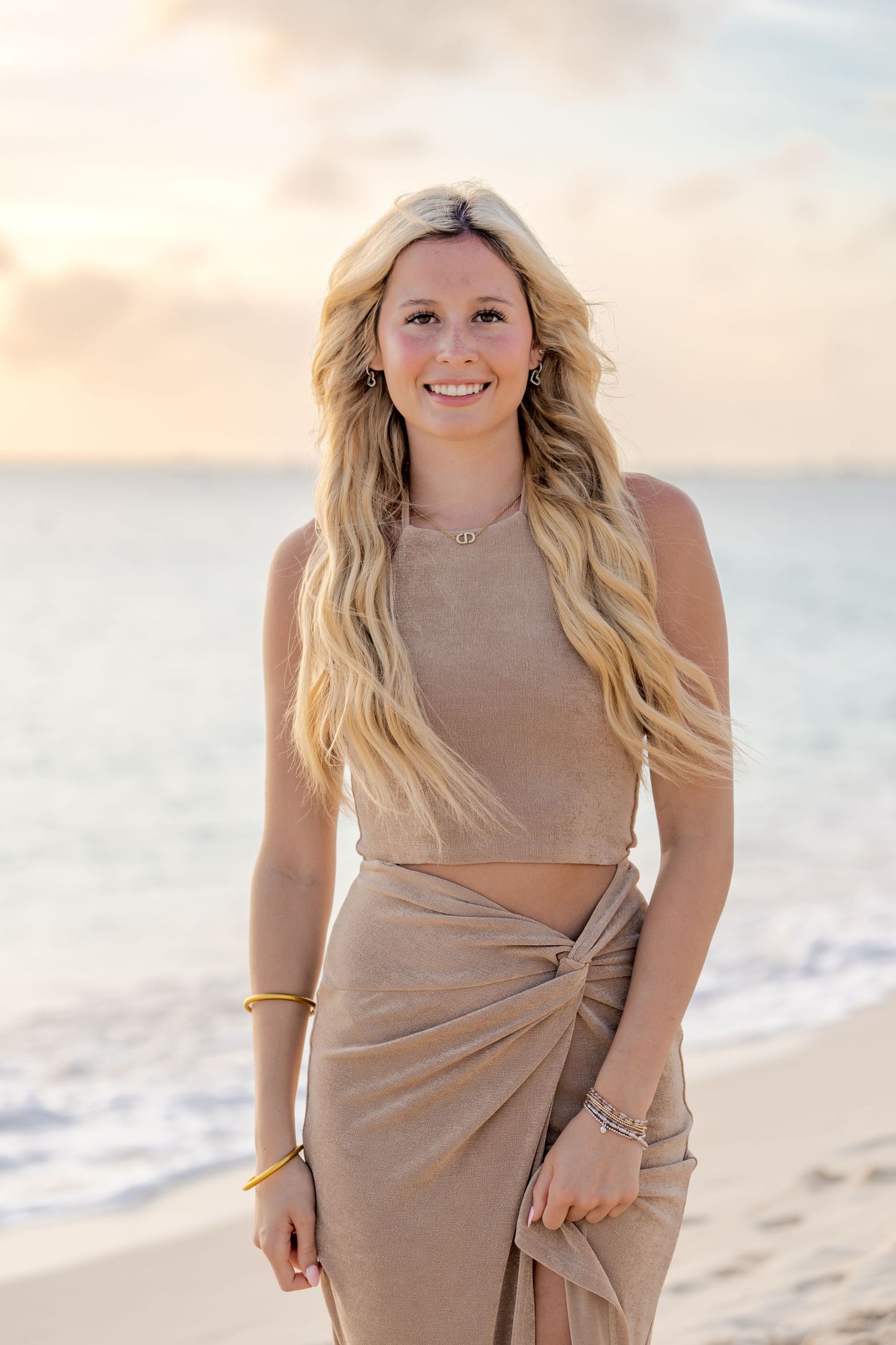 Woman in beige two-piece outfit poses on beach at sunset.