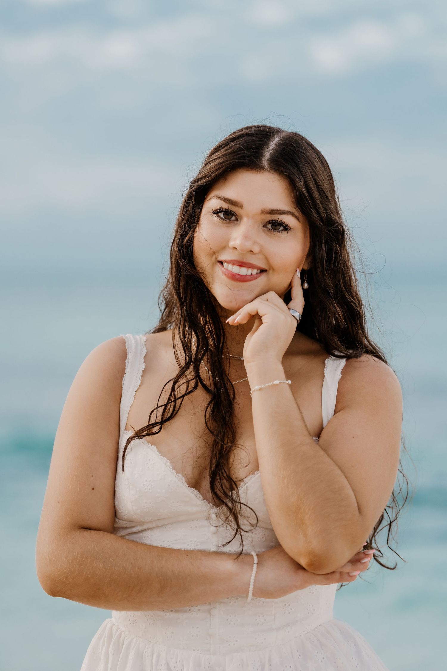 Woman in white dress poses playfully against blue ocean backdrop in beach photoshoot series.