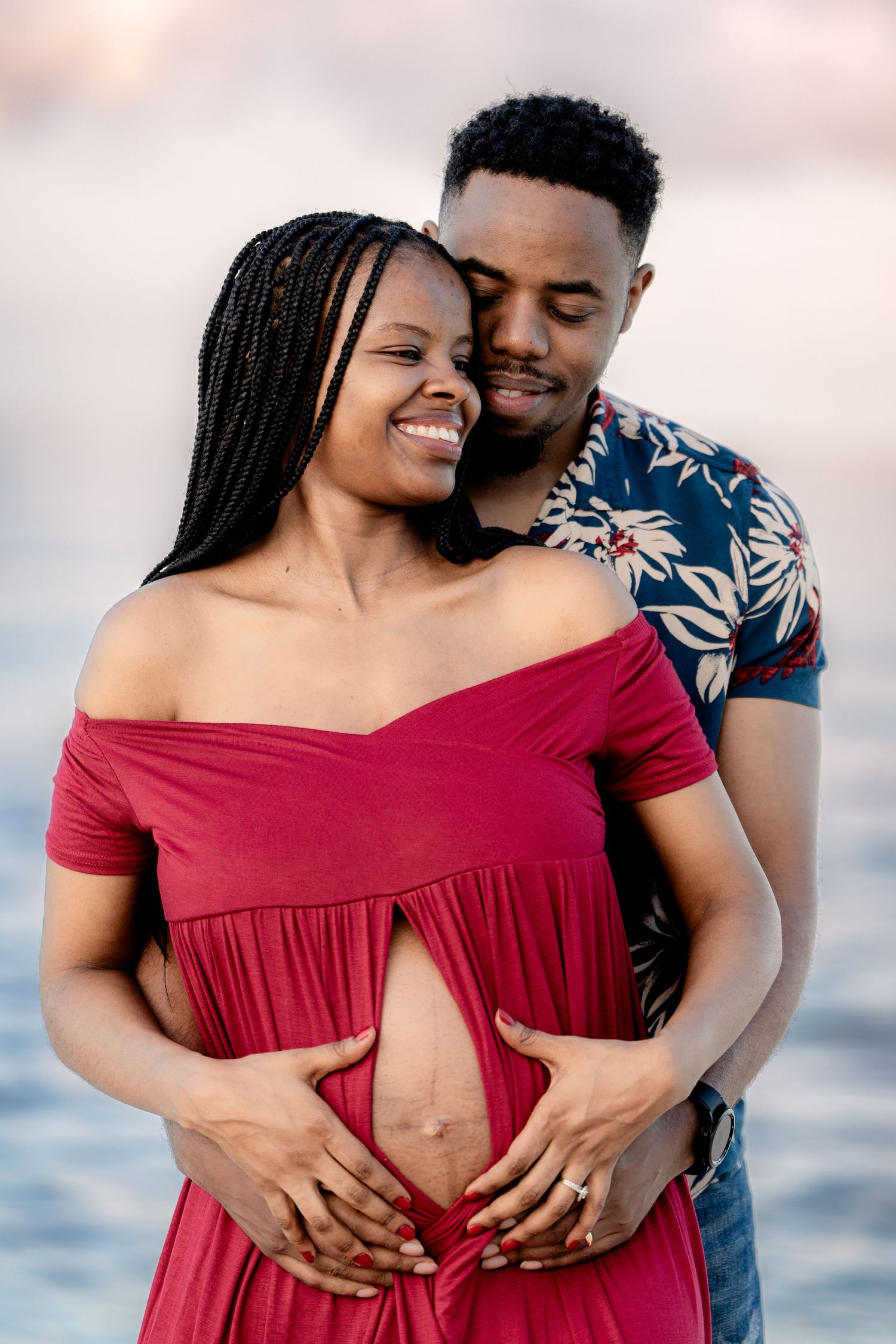 Couple shares intimate moment on beach wearing red dress and floral shirt during maternity session.