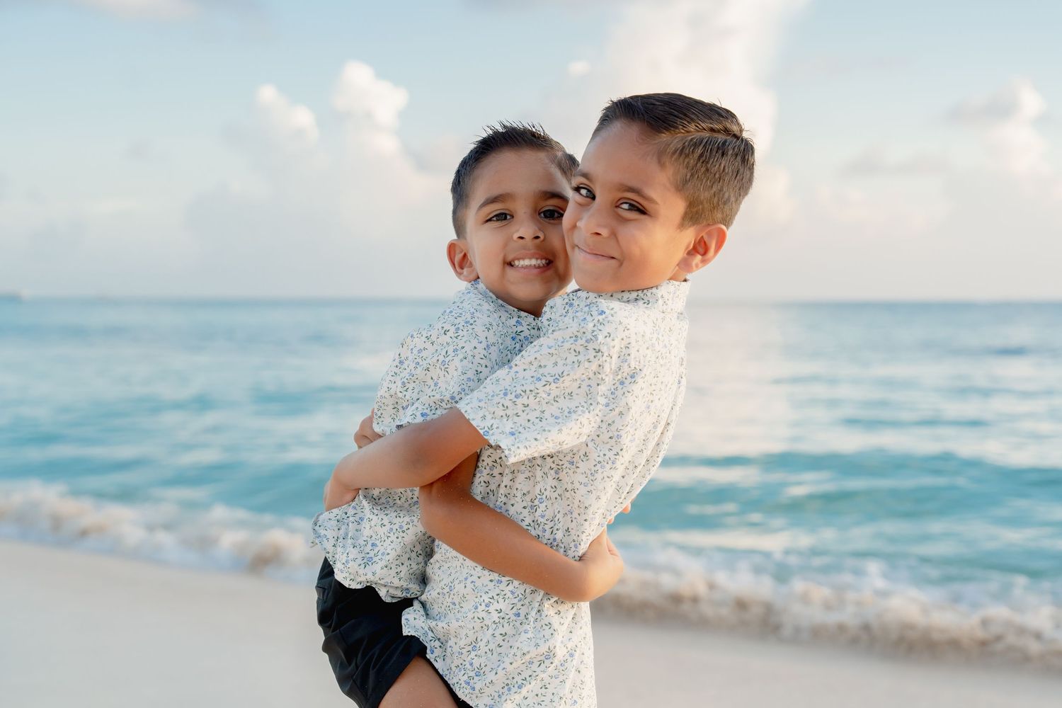 Two young boys hugging and smiling on a tropical beach with turquoise ocean waves in the background.