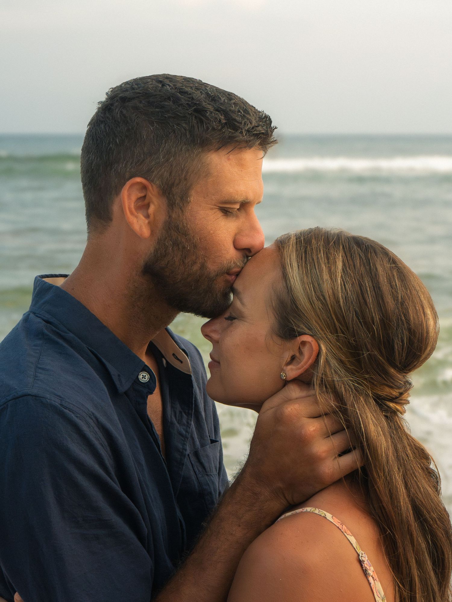 Romantic couple sharing a kiss on a beach with ocean waves visible behind them at dusk.