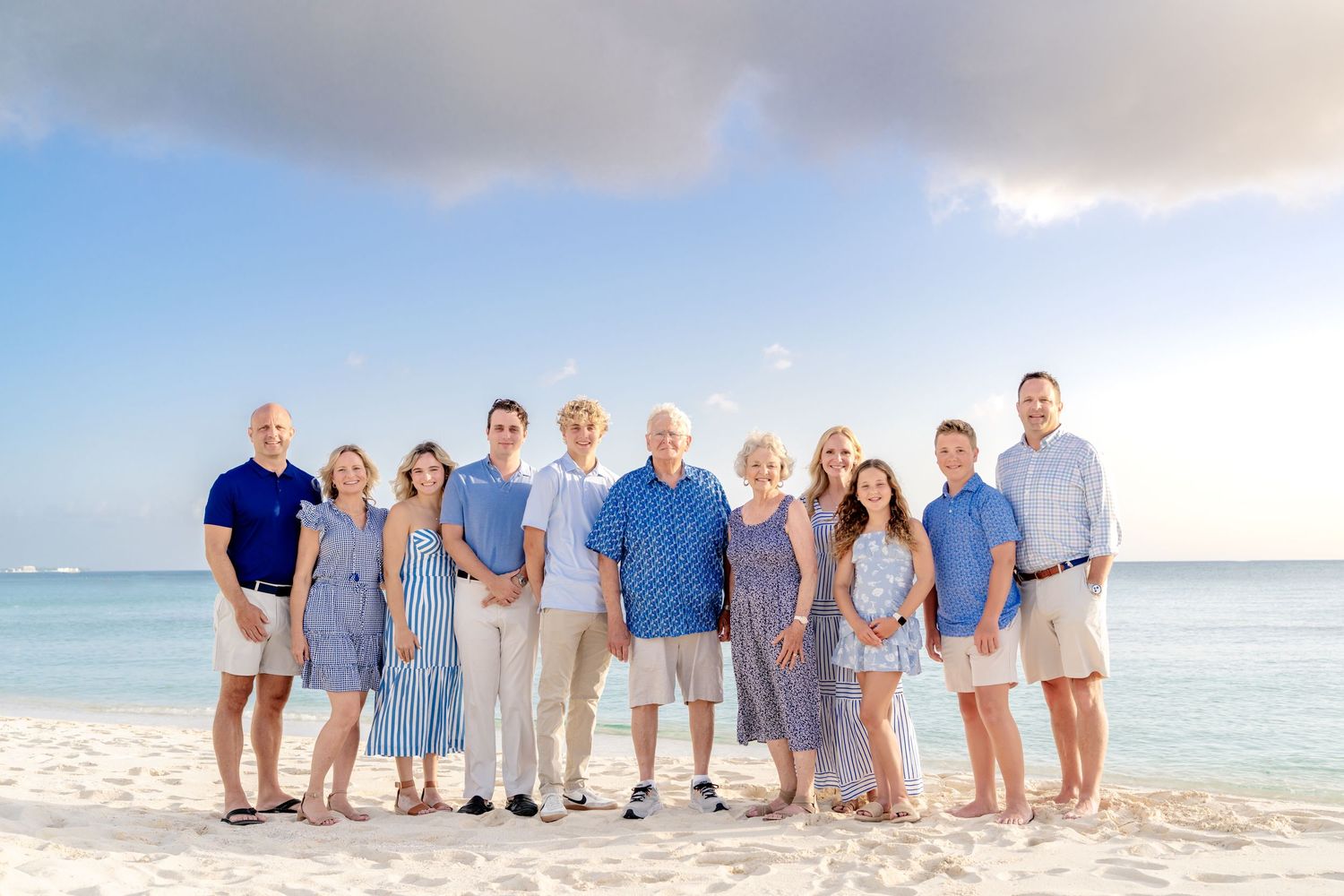 Multi-generational group in coordinated blue and white outfits poses on beach at sunset.