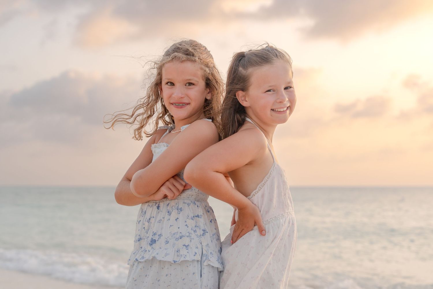 Two young girls hugging and smiling on a beach at sunset, wearing light summer dresses with ocean waves behind them.