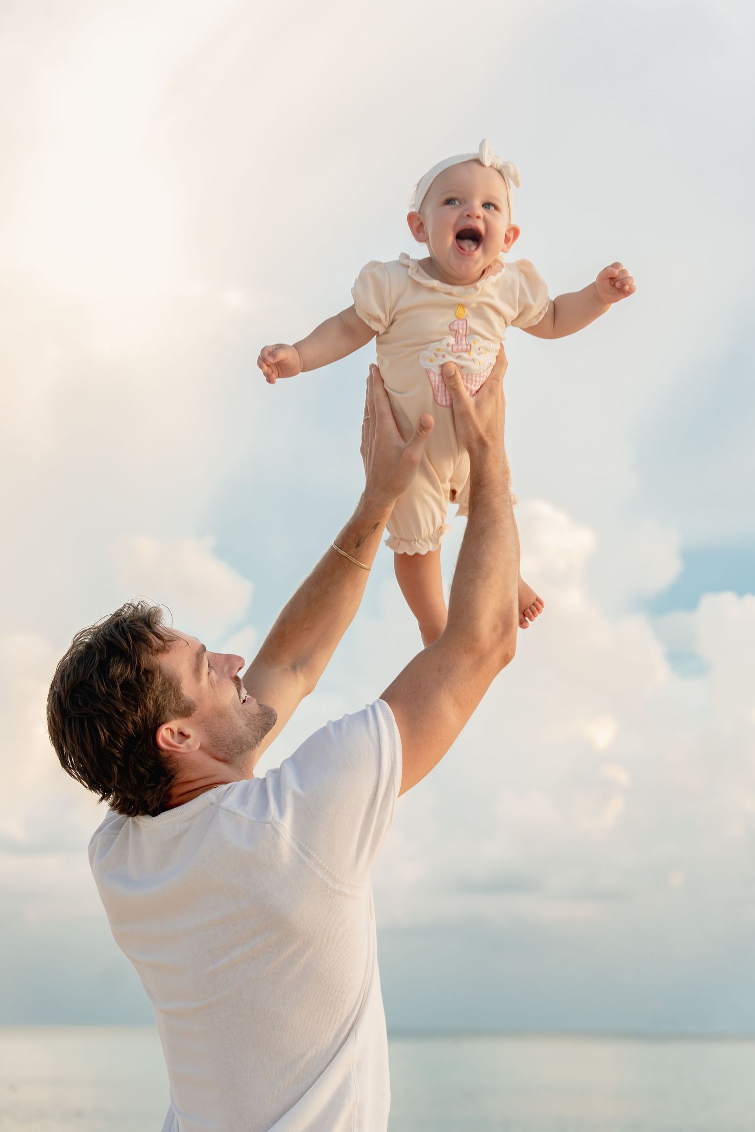 Father joyfully lifting laughing baby girl high in the air on a sunny beach with blue sky background.
