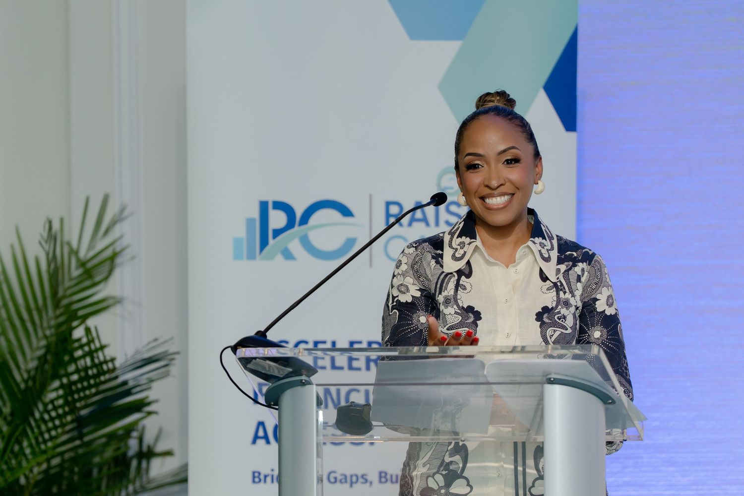 Presenter delivers speech at corporate podium with professional backdrop and palm plants.