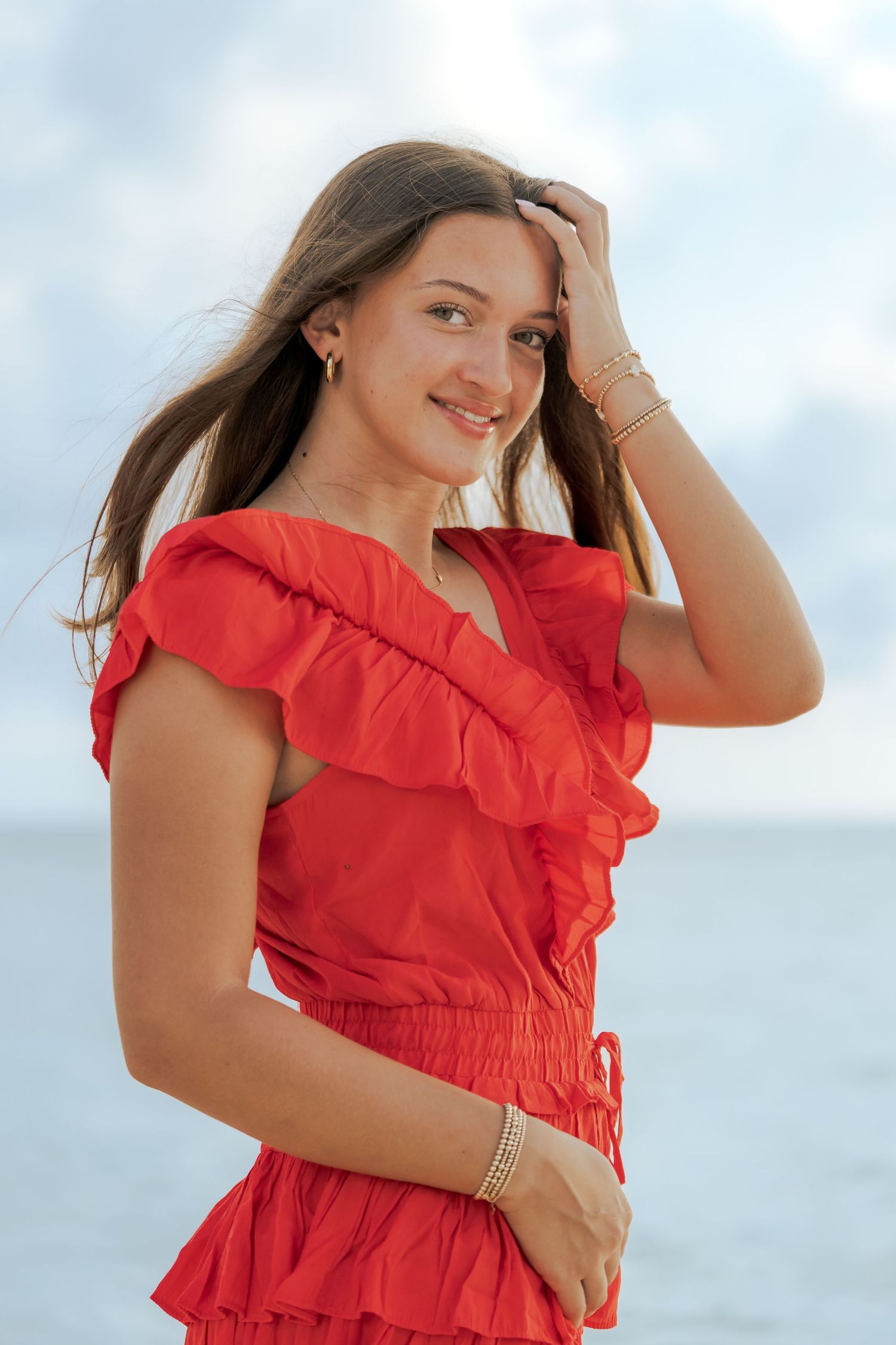 Woman in red ruffled summer dress poses against cloudy sky background at beach.