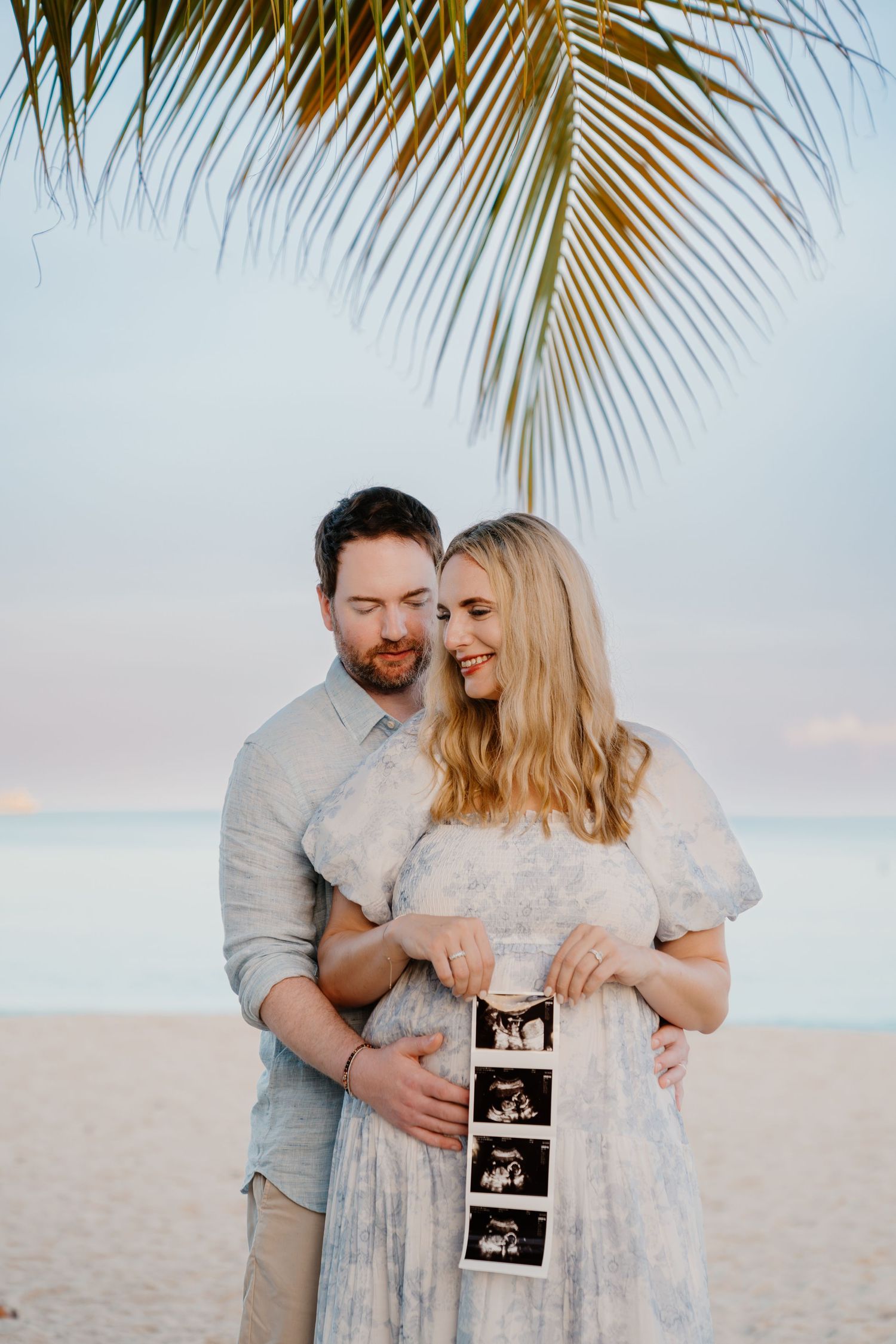 Beach pregnancy announcement photoshoot under palm trees with ultrasound images.