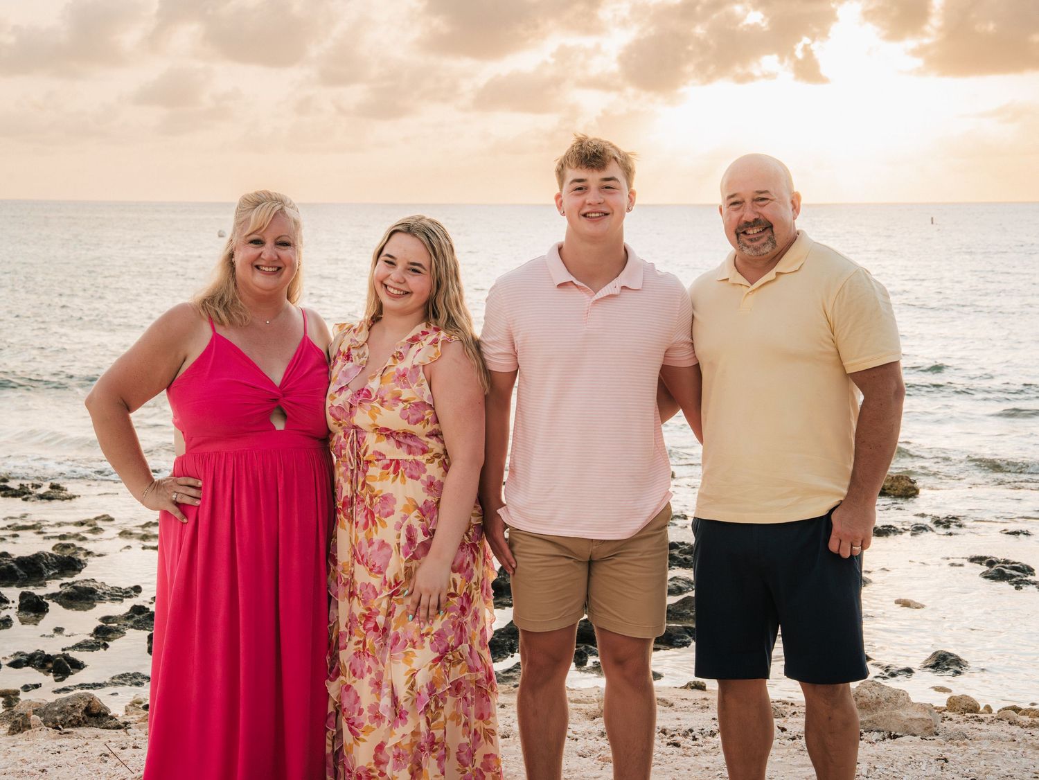 Family of four smiling together on a tropical beach at sunset, wearing colorful summer outfits.