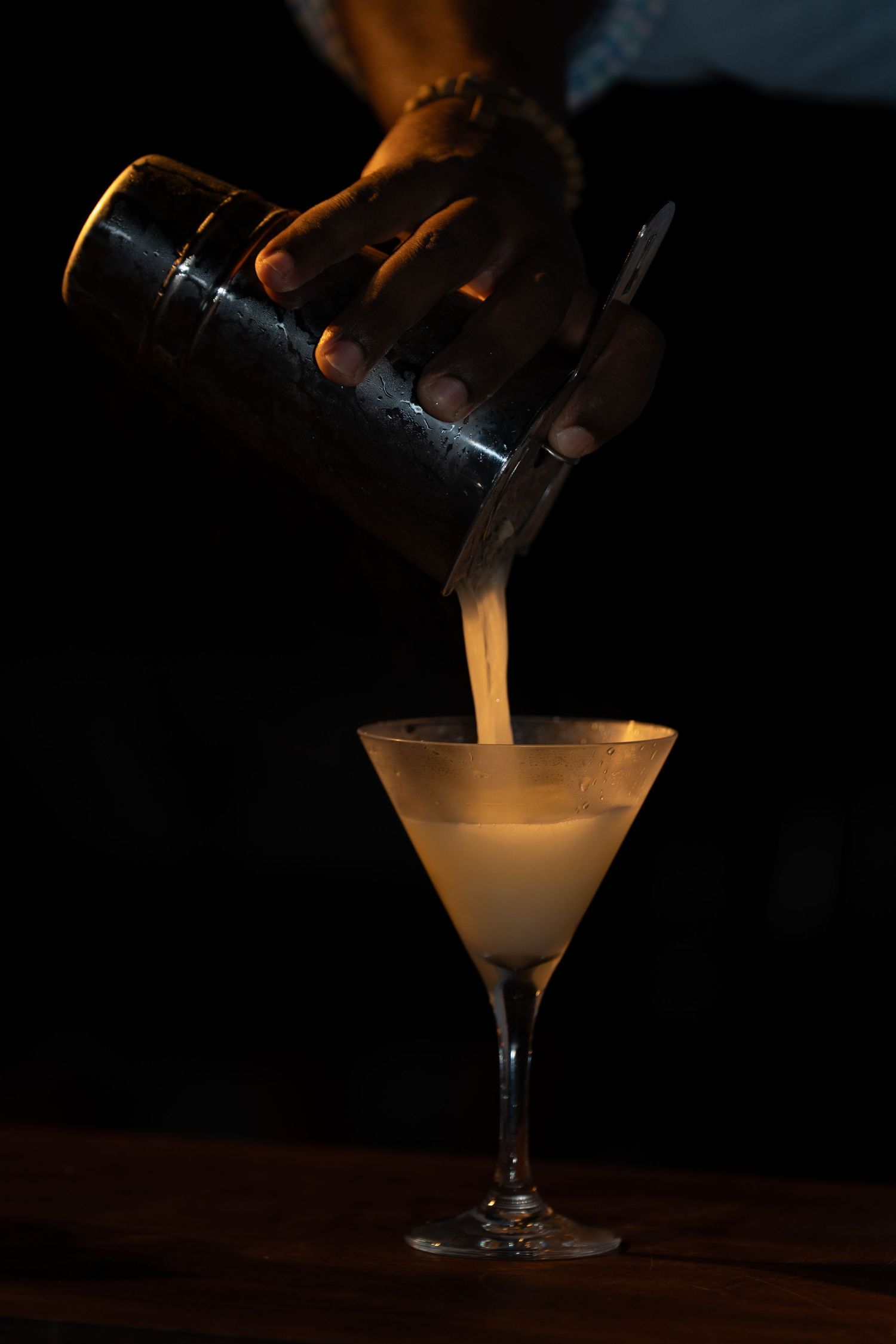 Bartender pouring a creamy cocktail into a martini glass in dark lighting.