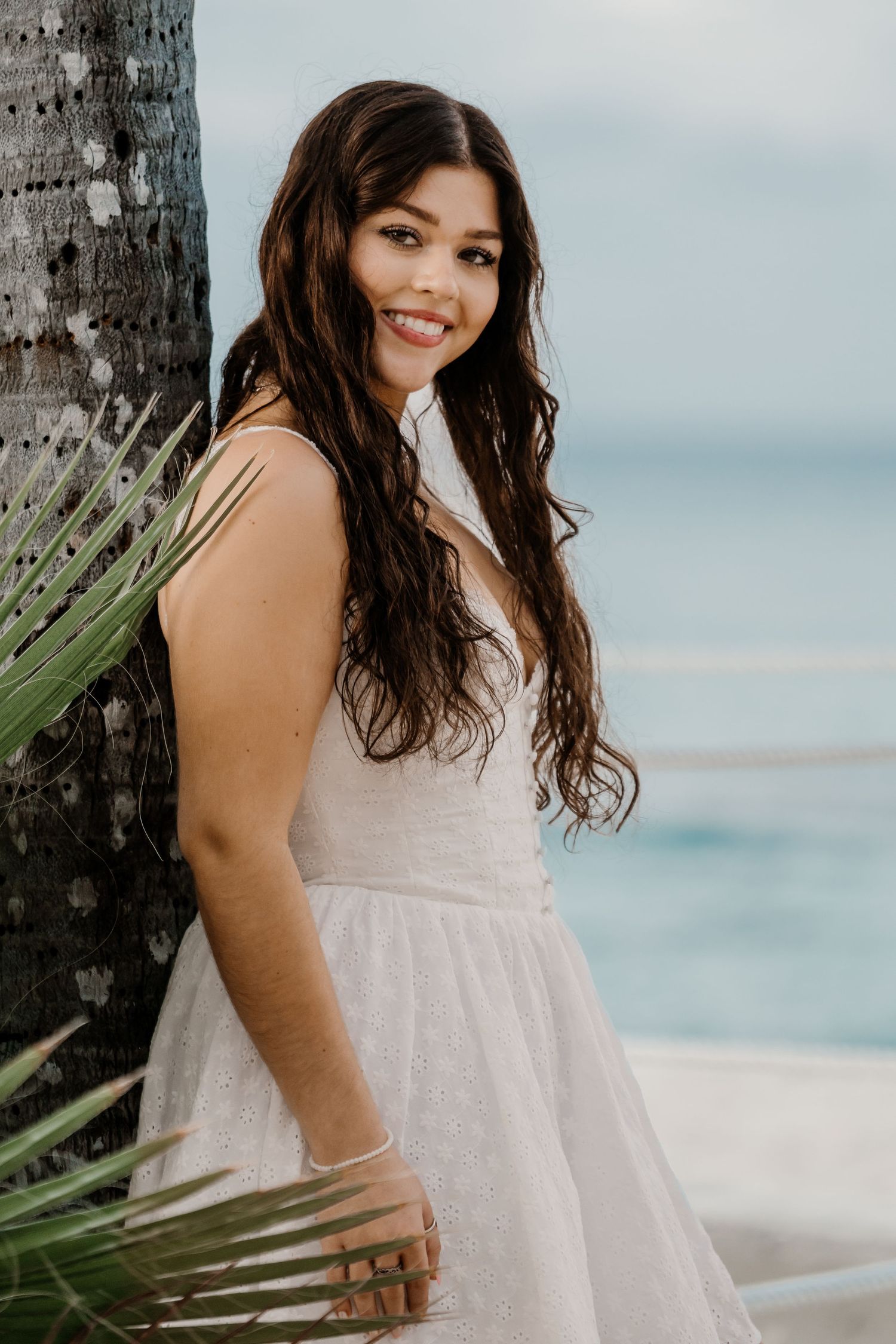 Tropical beach photoshoot featuring white dress and palm trees with ocean in background.