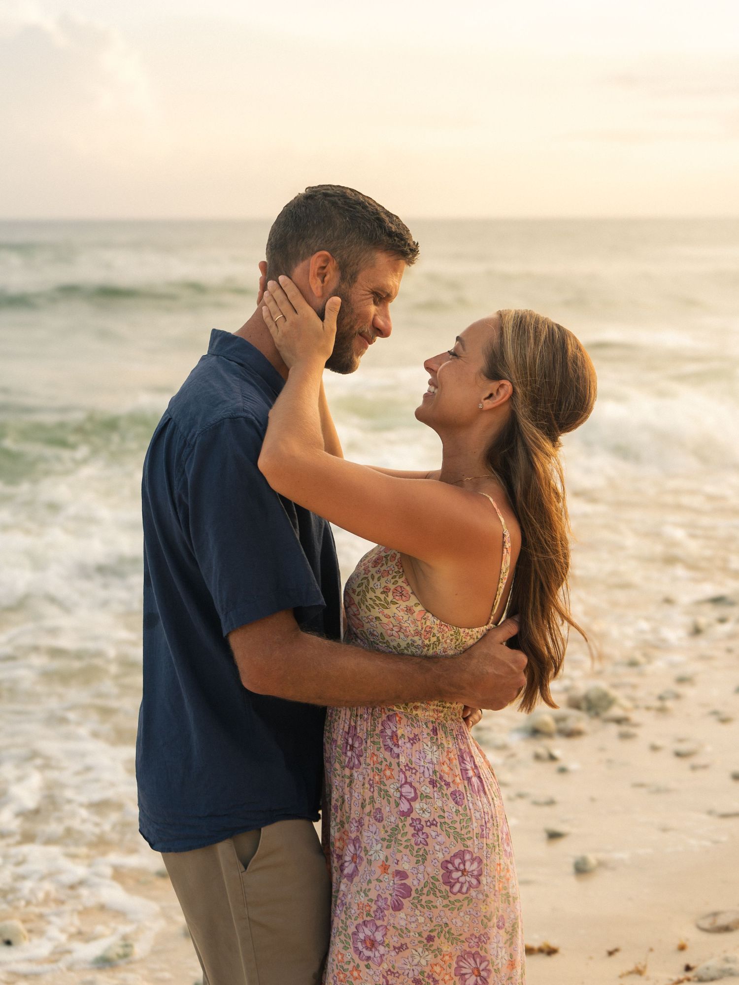 Couple embracing and smiling at each other on a beach at sunset with ocean waves in the background.