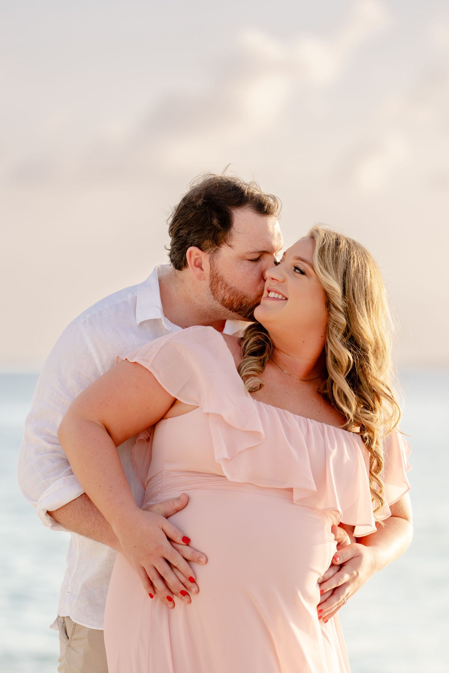 Romantic couple embraces in soft lighting wearing pink and white outfits during a beach photoshoot at sunset.
