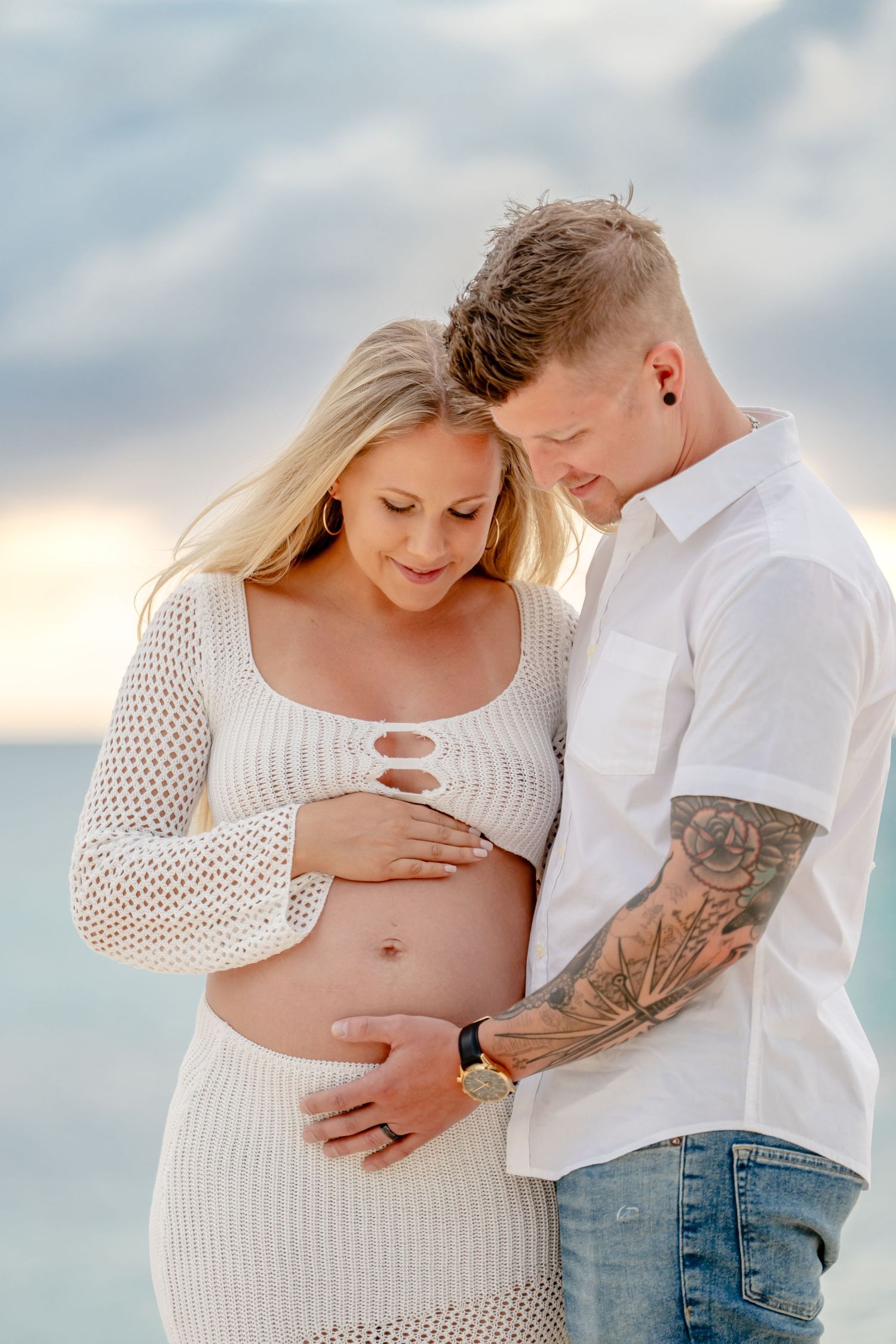 Intimate beach maternity photoshoot with couple in white outfits against cloudy sky.