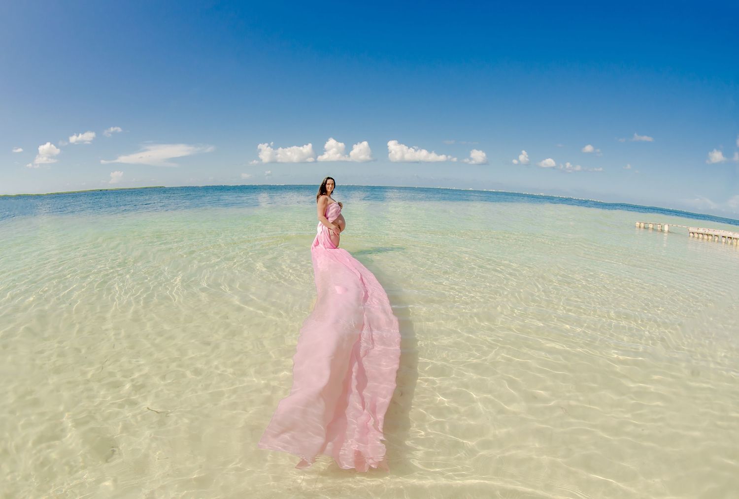 Long-distance beach landscape with person in flowing pink dress walking on shallow waters.