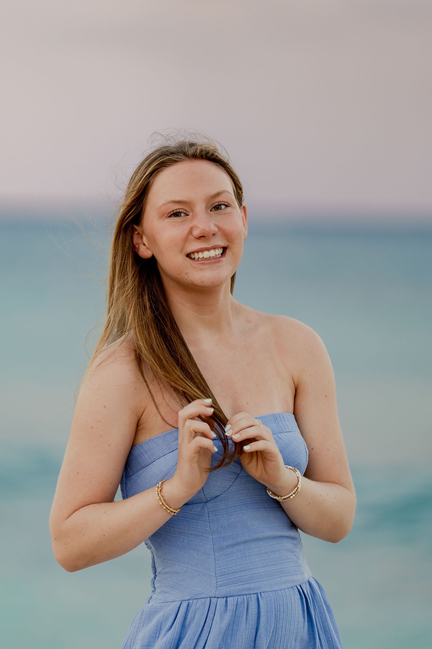 Woman in light blue sundress poses playfully on beach with ocean backdrop.