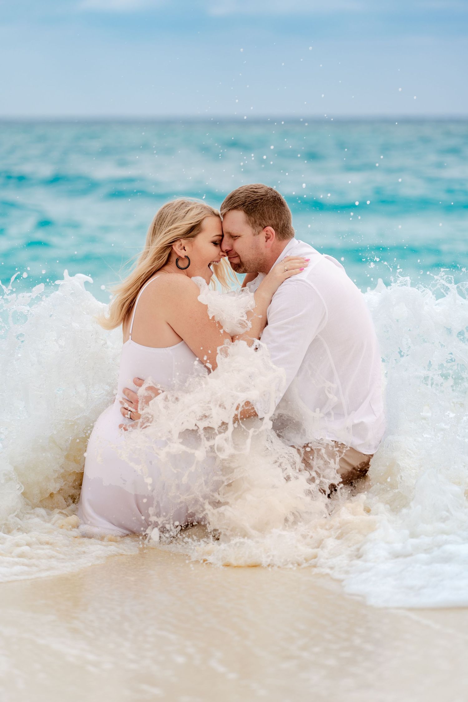 A splashing wave crashes around a couple embracing in white attire on a tropical beach.