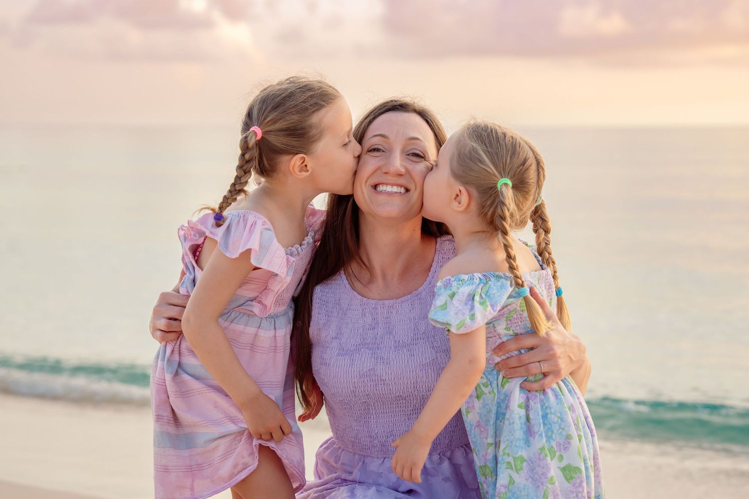 A smiling woman in a purple dress receives kisses from two young girls with braids on a beach at sunset.