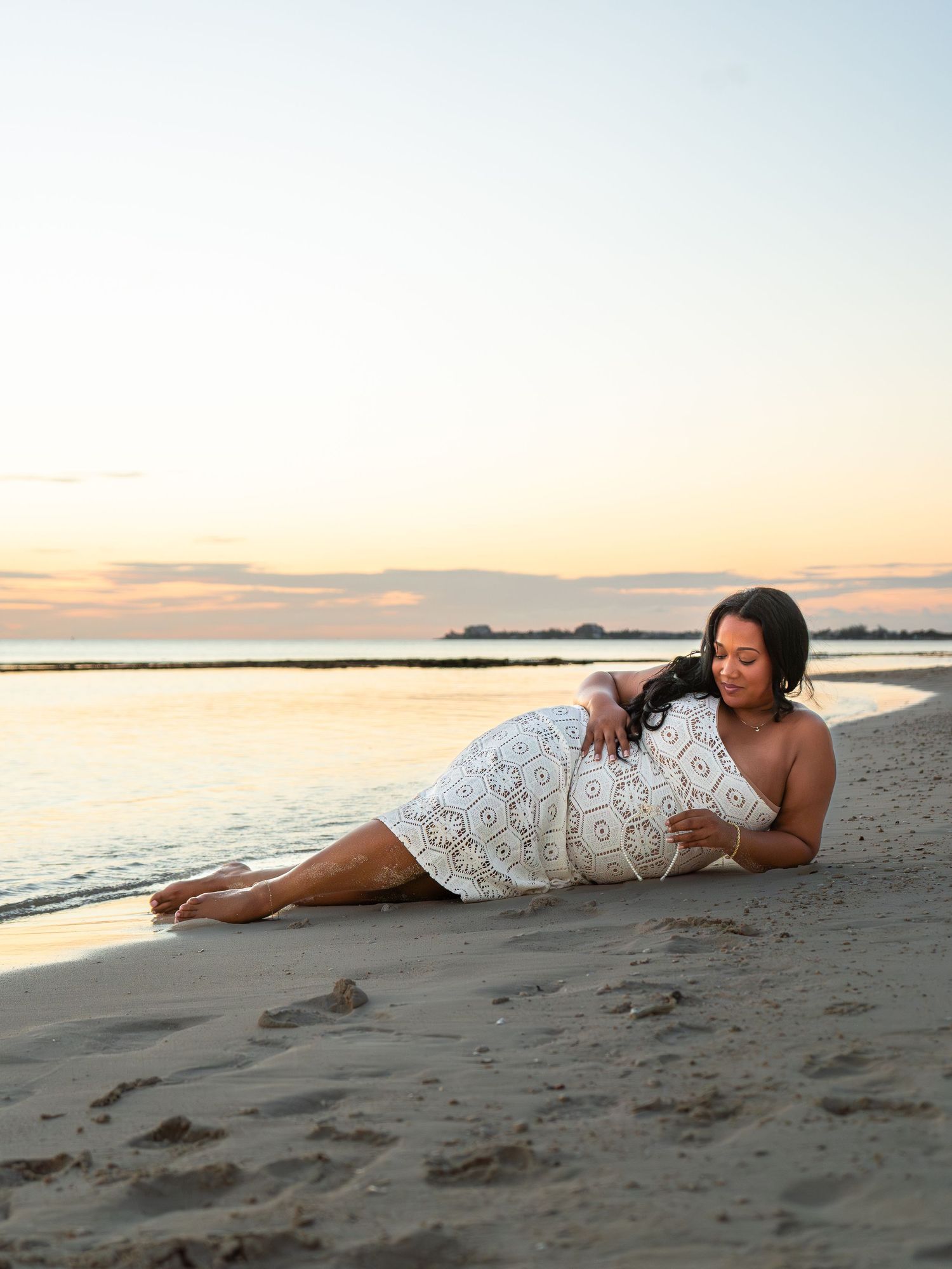 Pregnant woman in white lace dress reclining on sandy beach at golden sunset.