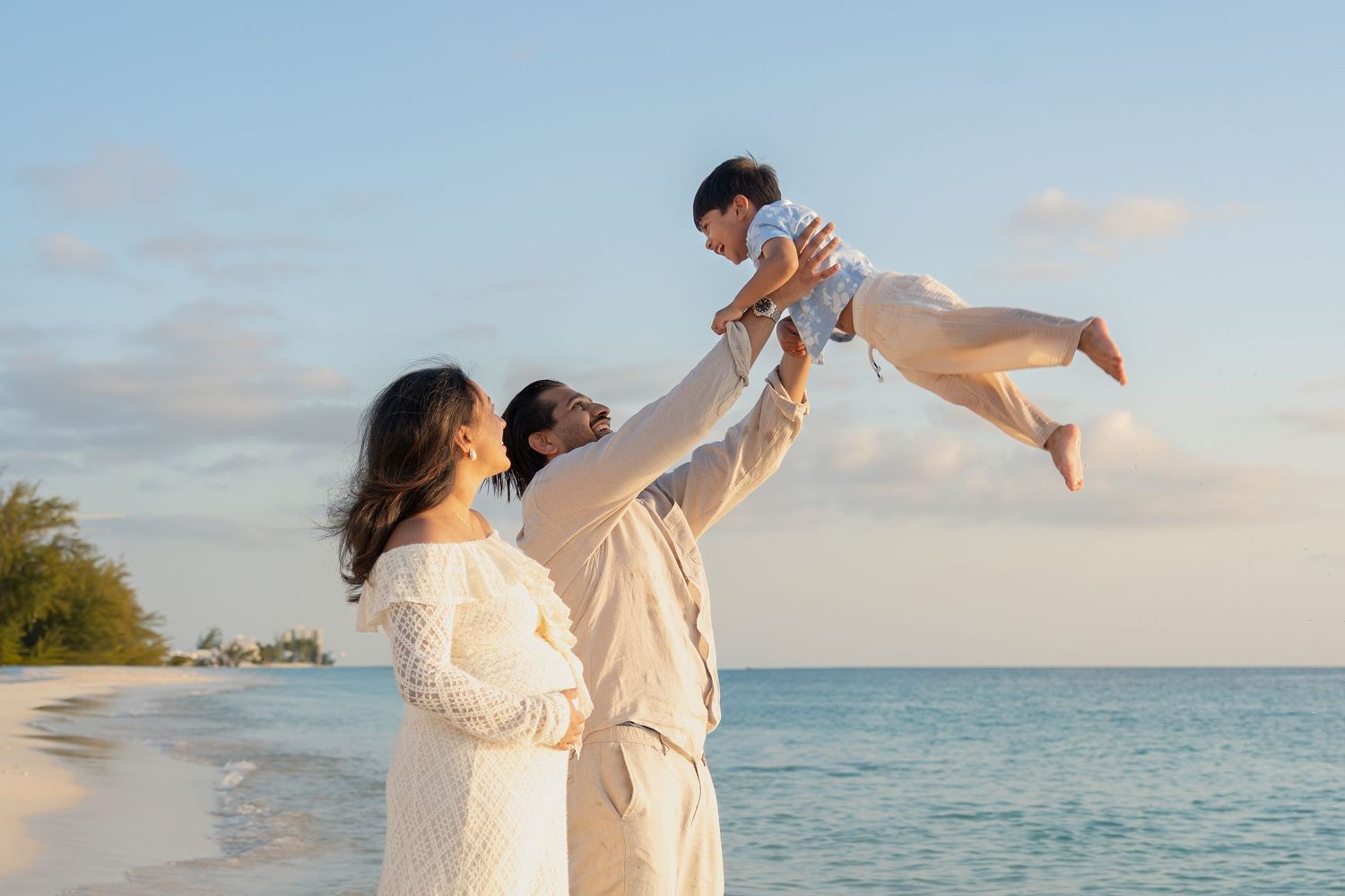 Family in white outfits on a beach, father lifting baby in the air while mother watches by the ocean.