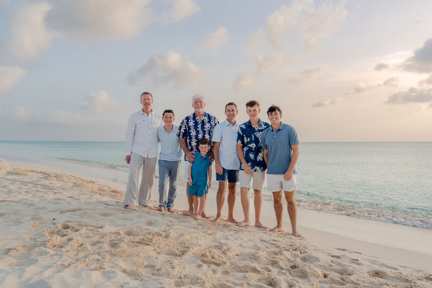 Multi-generational family of seven poses together on a white sand beach at sunset with turquoise ocean waters behind them.