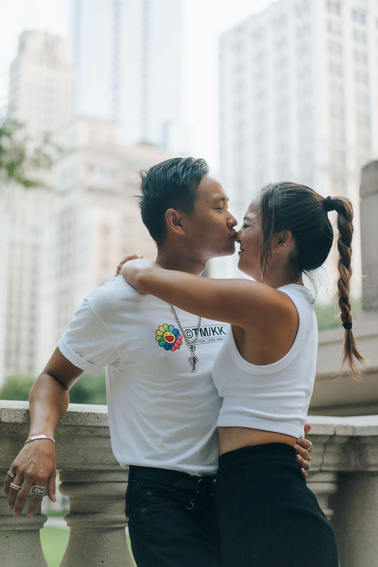 Couple sharing a romantic kiss outdoors in an urban setting, wearing matching white t-shirts with city buildings in the background.