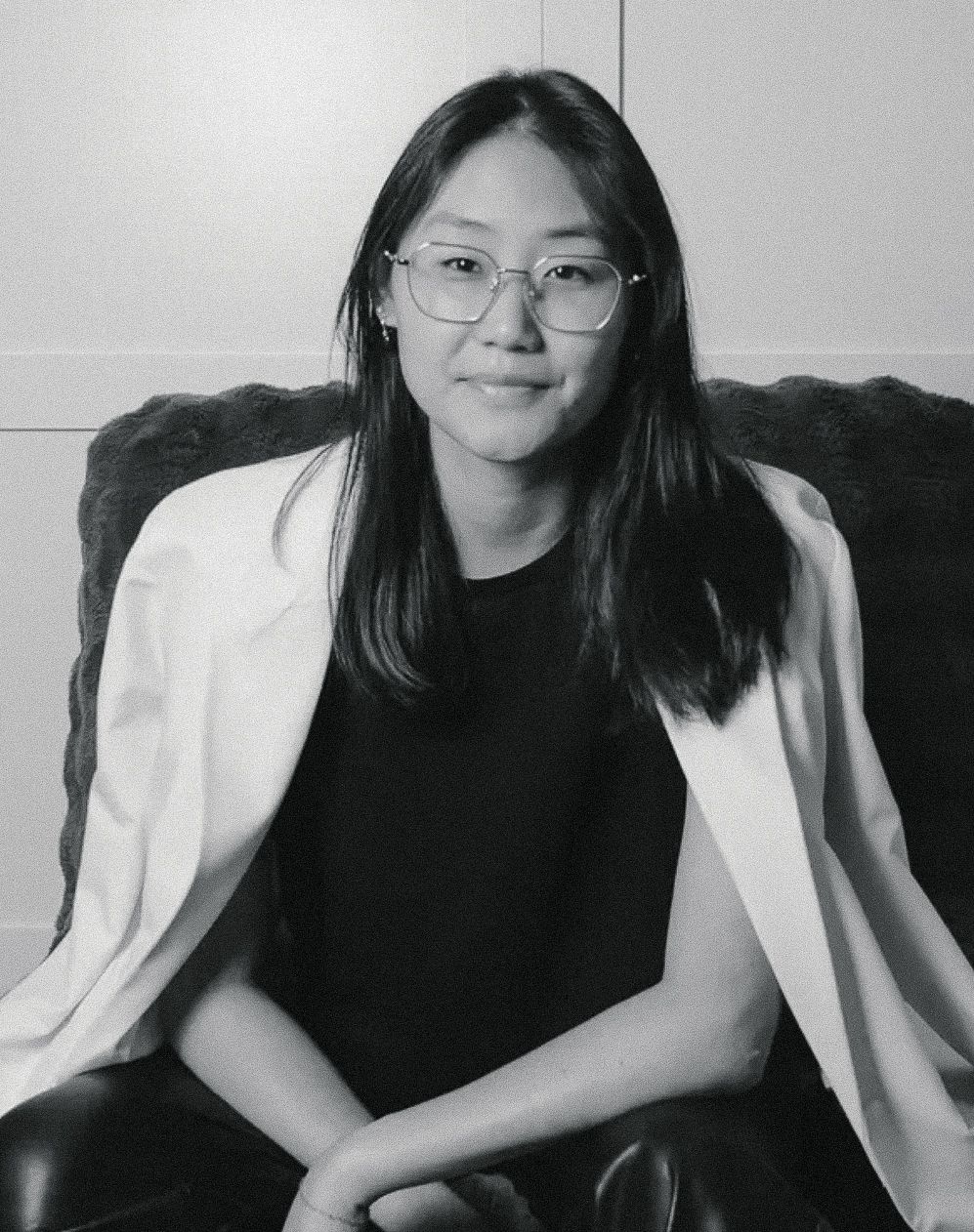 A young woman with glasses and long dark hair smiles, wearing a white blazer over a black top, seated in a chair.